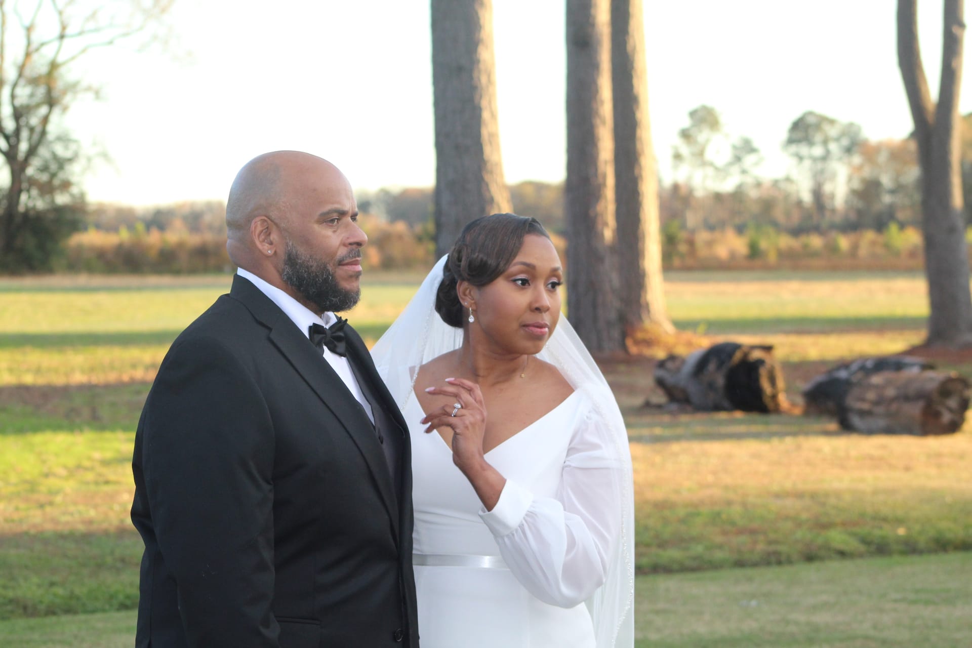 Bride in a long-sleeve white gown and veil stands beside her tuxedo-clad groom on the open farmland fields of Scarborough House estate during golden-hour light.