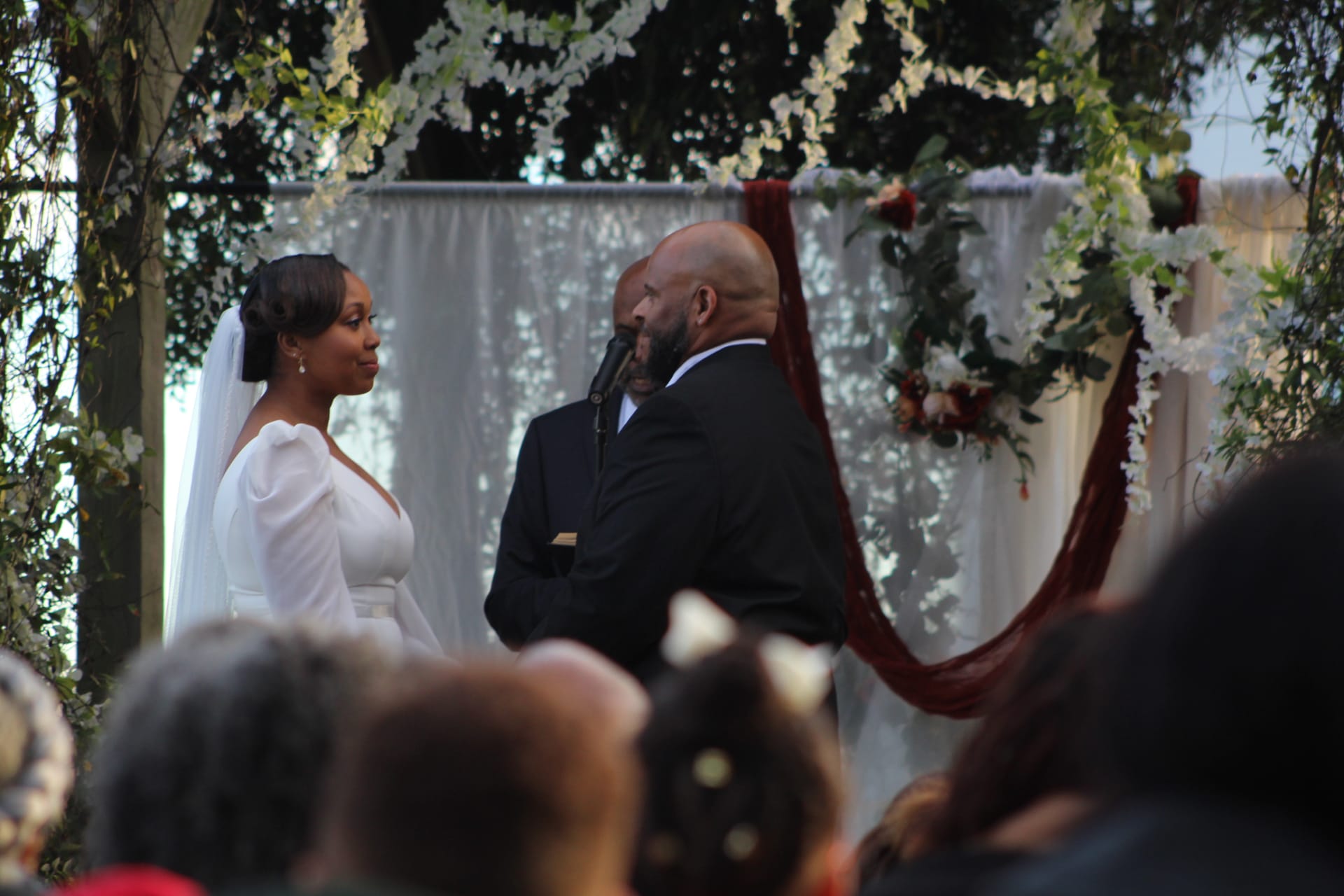 Bride in a white gown and veil faces her groom beneath a lush floral arch draped with white flowers and greenery during an outdoor wedding ceremony at Scarborough House.
