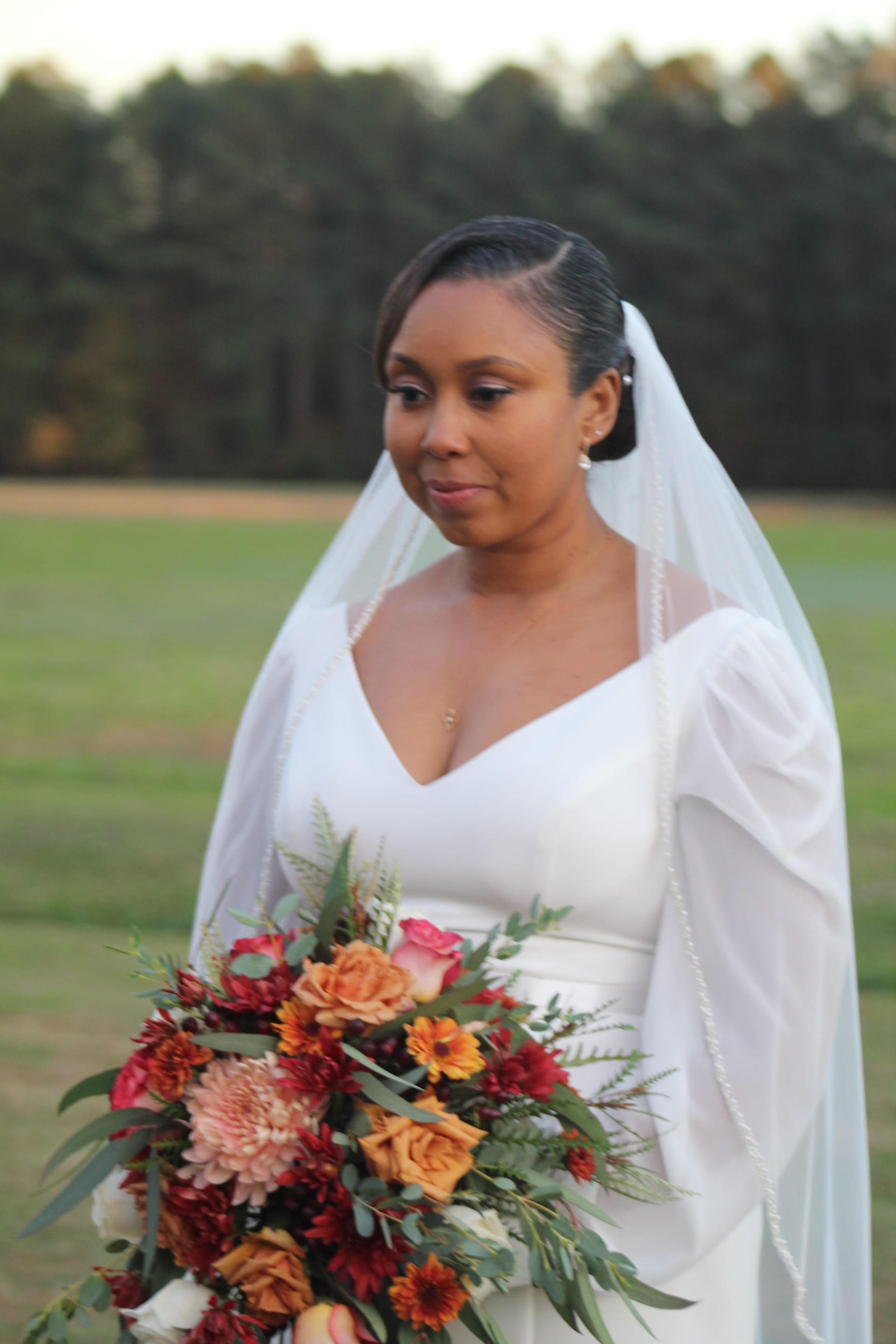 Bride in a white gown and veil holding a lush autumn bouquet of burgundy, orange, and blush flowers stands in an open field at golden hour with trees in the background at Scarborough House.