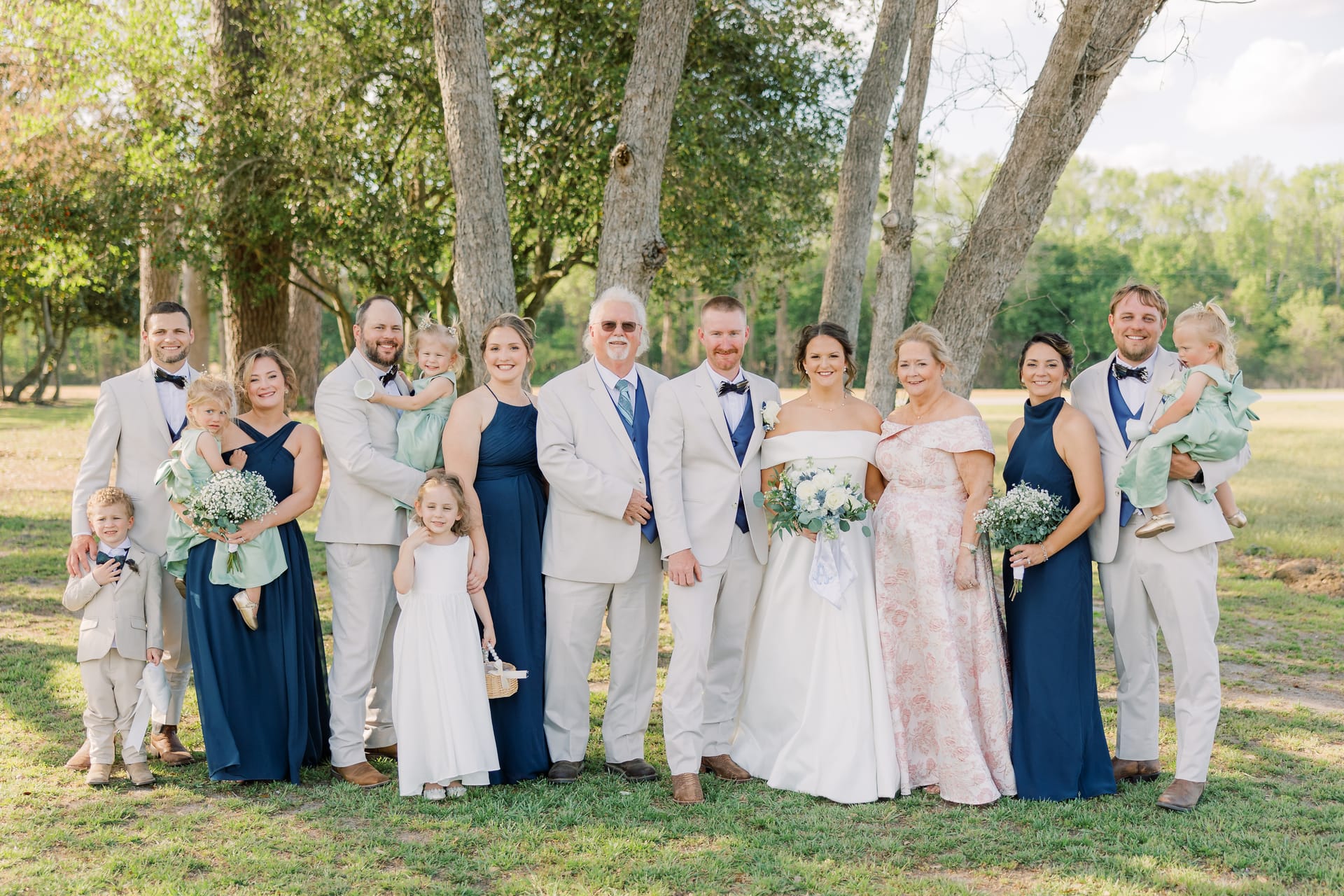 Large wedding party including bride and groom, groomsmen in tan suits, bridesmaids in navy gowns, and young children posing together beneath tall oak trees on the grounds of Scarborough House.