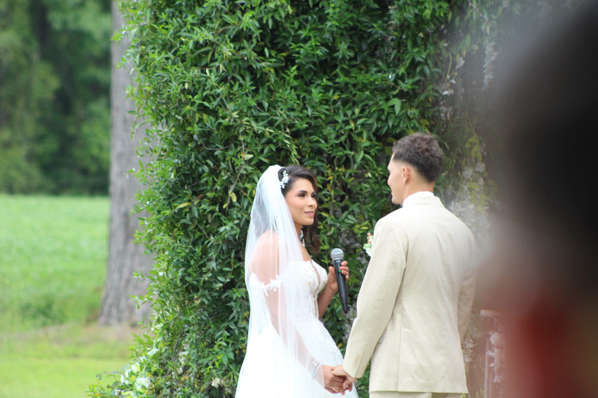Bride in a white lace gown and veil holds a microphone while reciting vows to her groom in a tan suit against a lush ivy-covered backdrop at an outdoor garden ceremony.