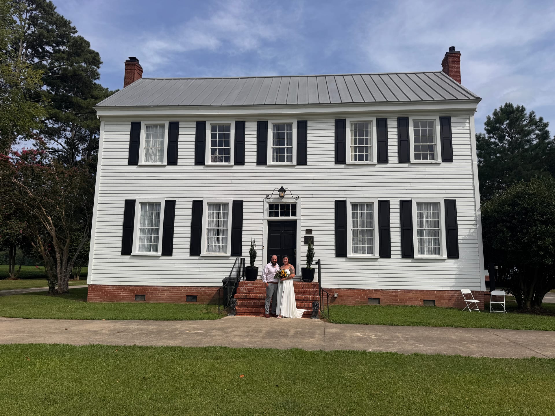 Bride and groom pose together on the brick front steps of the white 1821 Scarborough House manor, flanked by black shuttered windows and surrounded by lush green lawns under a partly cloudy sky.