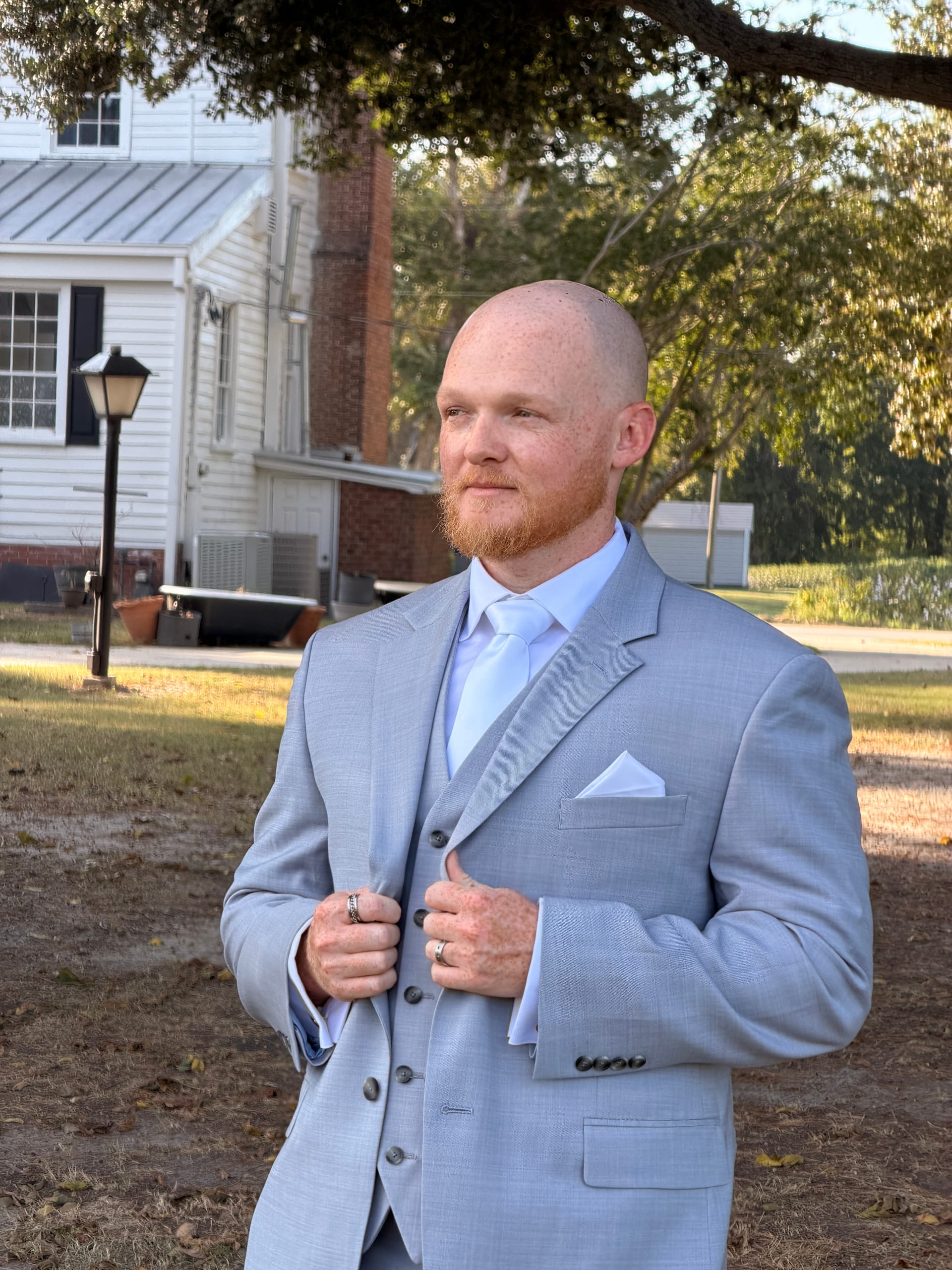 Dapper groom in a light gray three-piece suit and white tie adjusts his lapel while standing on the grounds of the historic Scarborough House estate.