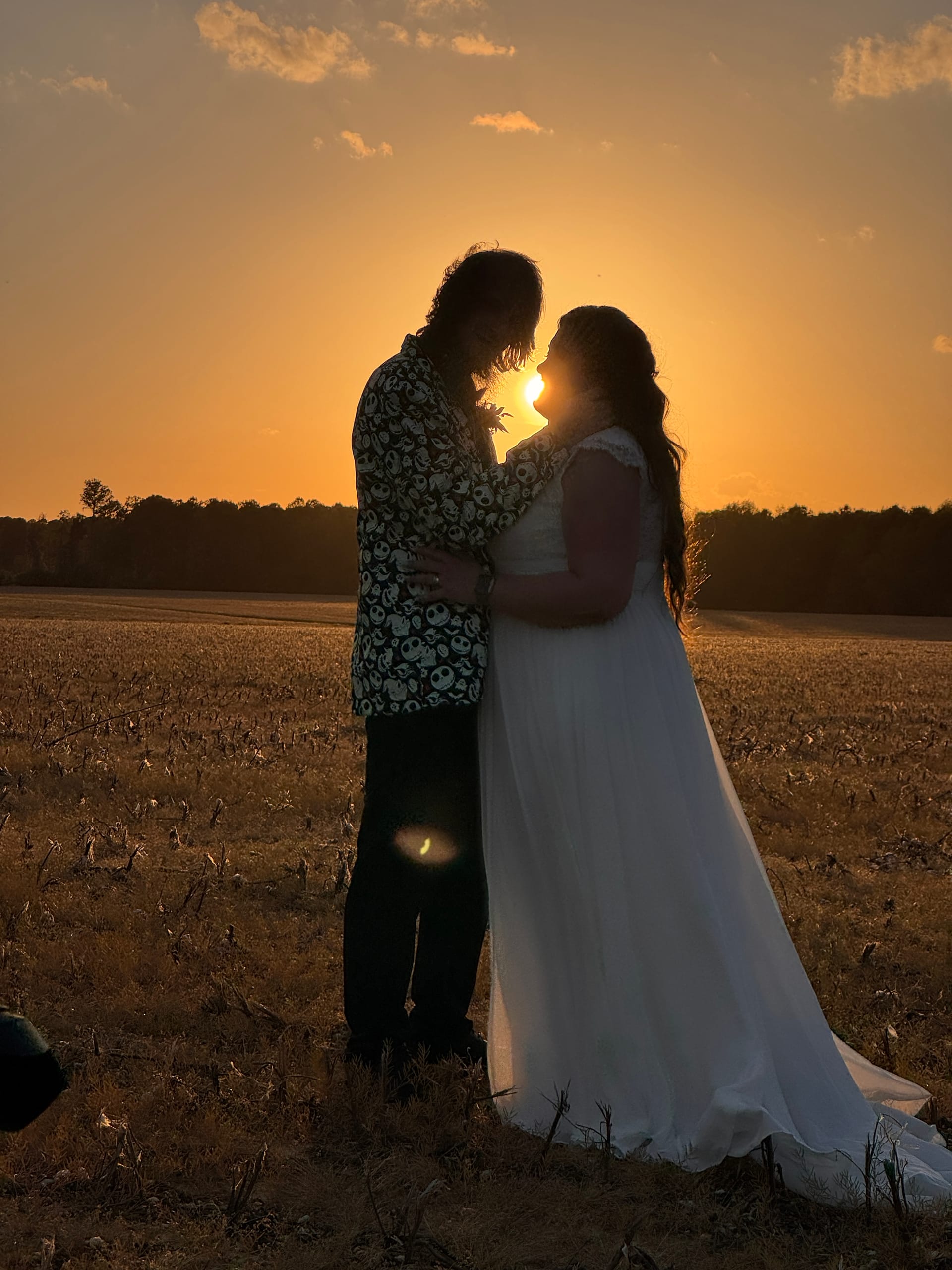 Bride in a flowing white gown and groom in a patterned shirt share an intimate moment as silhouettes against a glowing golden sunset over open farmland fields at Scarborough House.