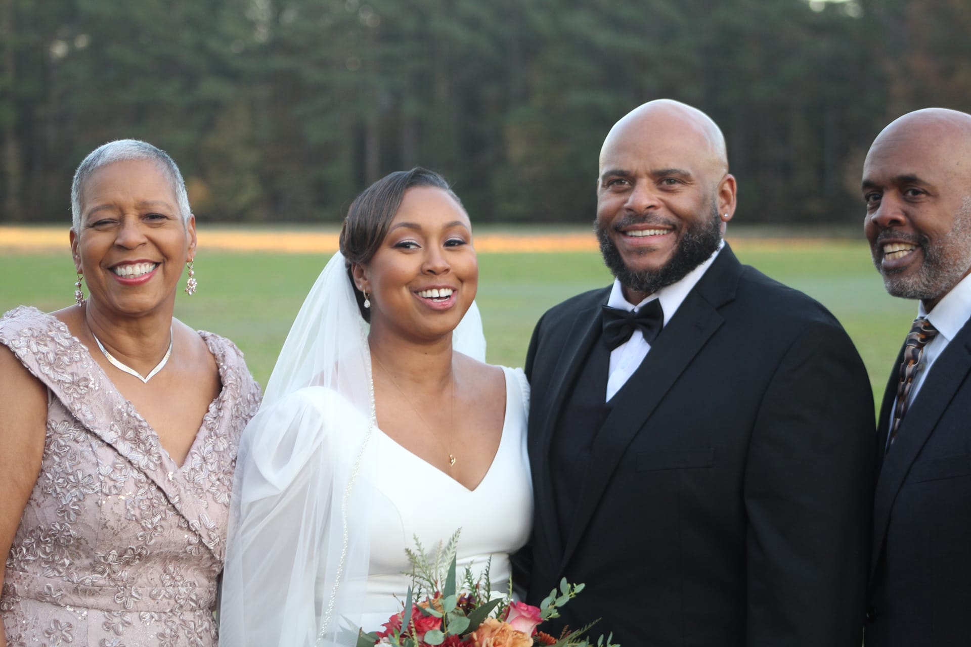 Bride in white gown and veil stands smiling with three family members including the groom in a black tuxedo against a soft tree-lined field backdrop at Scarborough House.