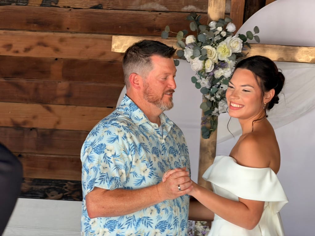 A smiling bride in an off-shoulder white gown holds hands with a man in a floral shirt beneath a circular floral arch adorned with white roses and eucalyptus at Scarborough House.