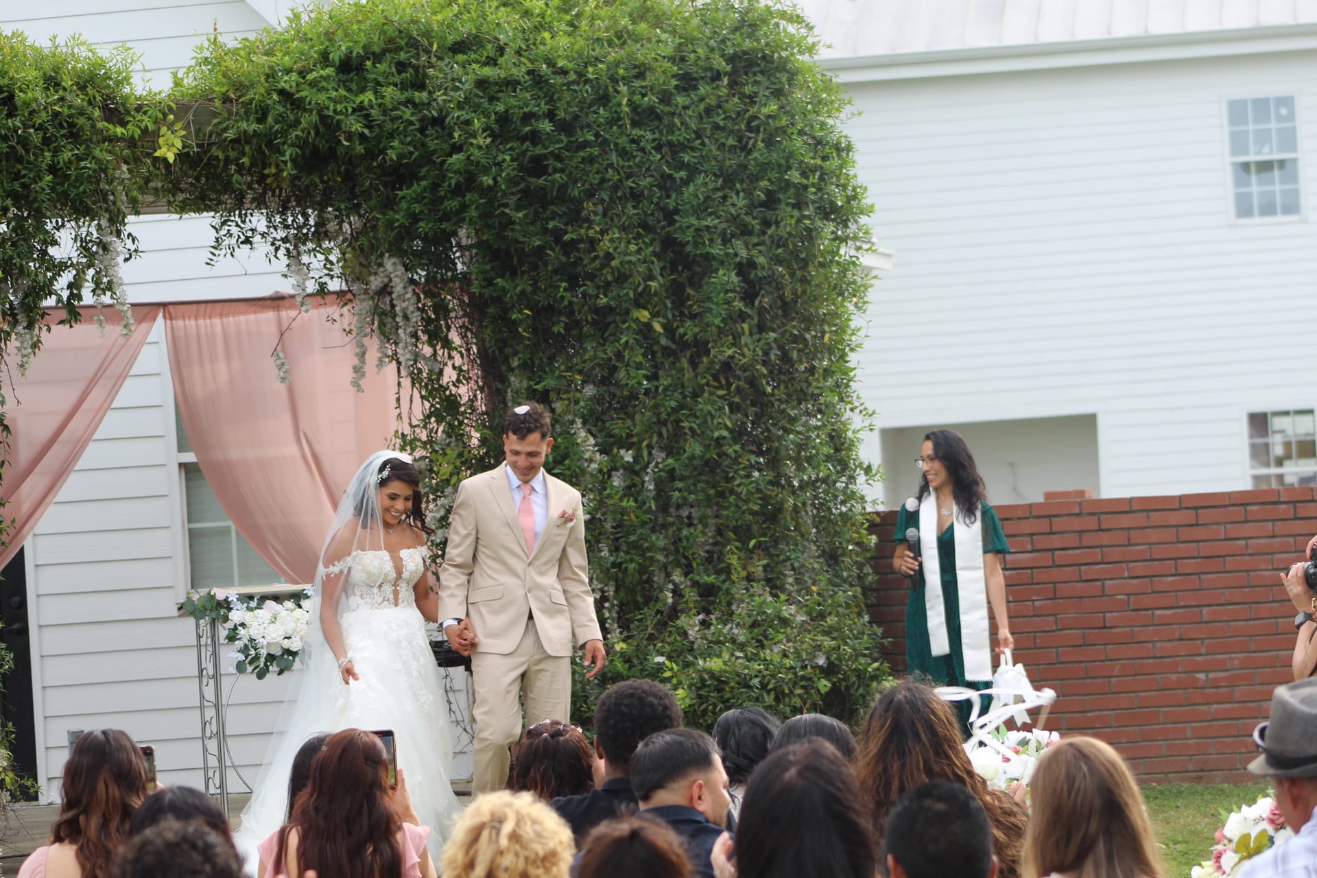 Bride in a lace ball gown and groom in a tan suit walk joyfully down the aisle during their outdoor wedding ceremony at Scarborough House, framed by a floral arch and lush greenery.