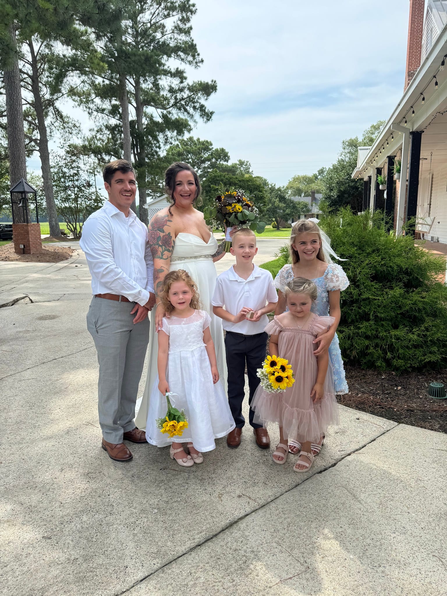Bride in a white gown holding a sunflower bouquet stands with her groom and four children dressed for the wedding celebration outside Scarborough House estate.