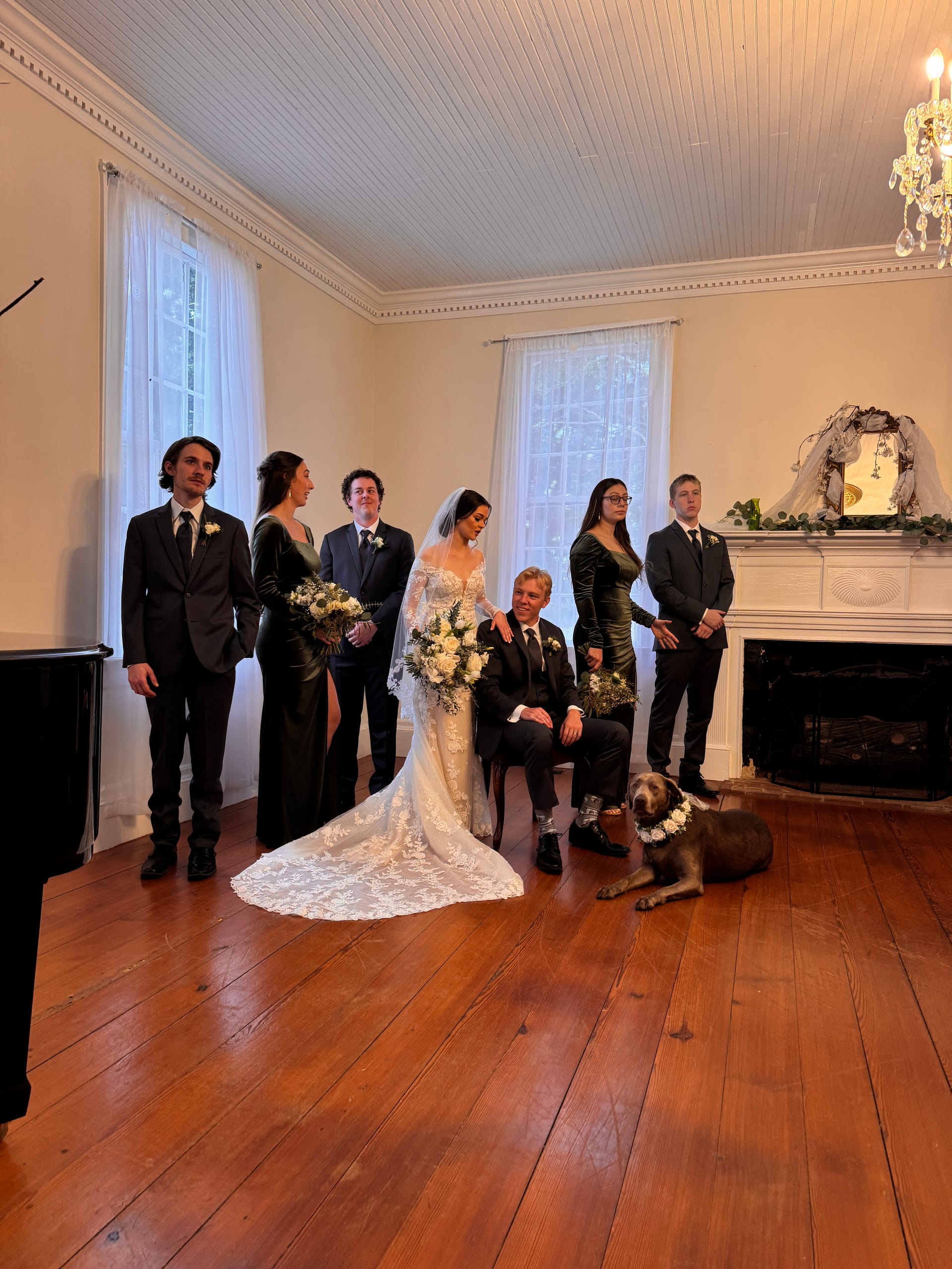Bride in a lace gown and groom in a black suit pose with their wedding party dressed in dark attire inside the historic grand hall of Scarborough House, with their dog wearing a floral collar lying at their feet.