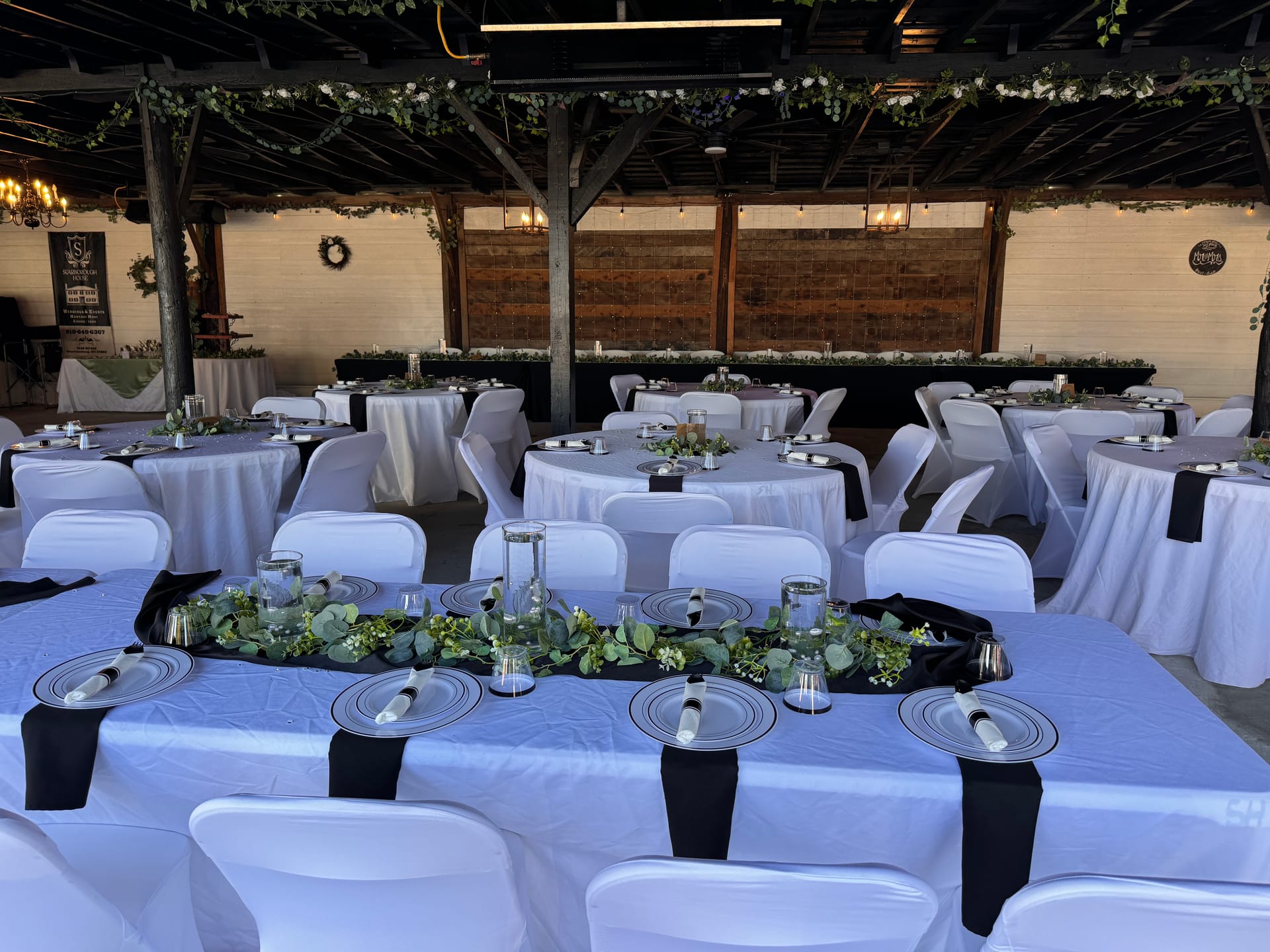 Elegantly set reception tables with white linens, black table runners, silver charger plates, and eucalyptus garland centerpieces under a rustic wood-beamed pavilion at Scarborough House.