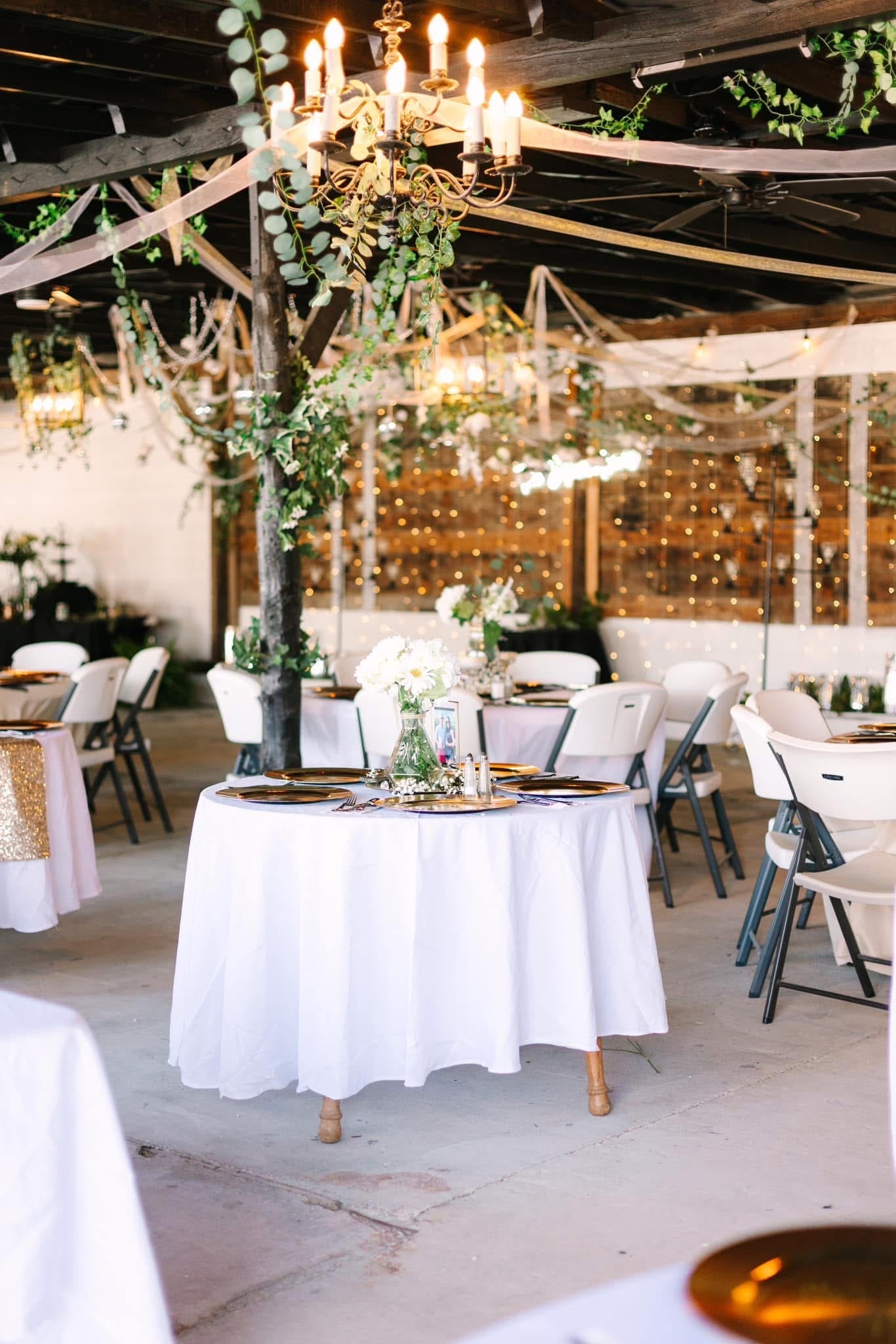 Elegantly set reception tables with white linens, gold charger plates, white floral centerpieces, and greenery-draped chandeliers inside a warmly lit rustic pavilion at Scarborough House.