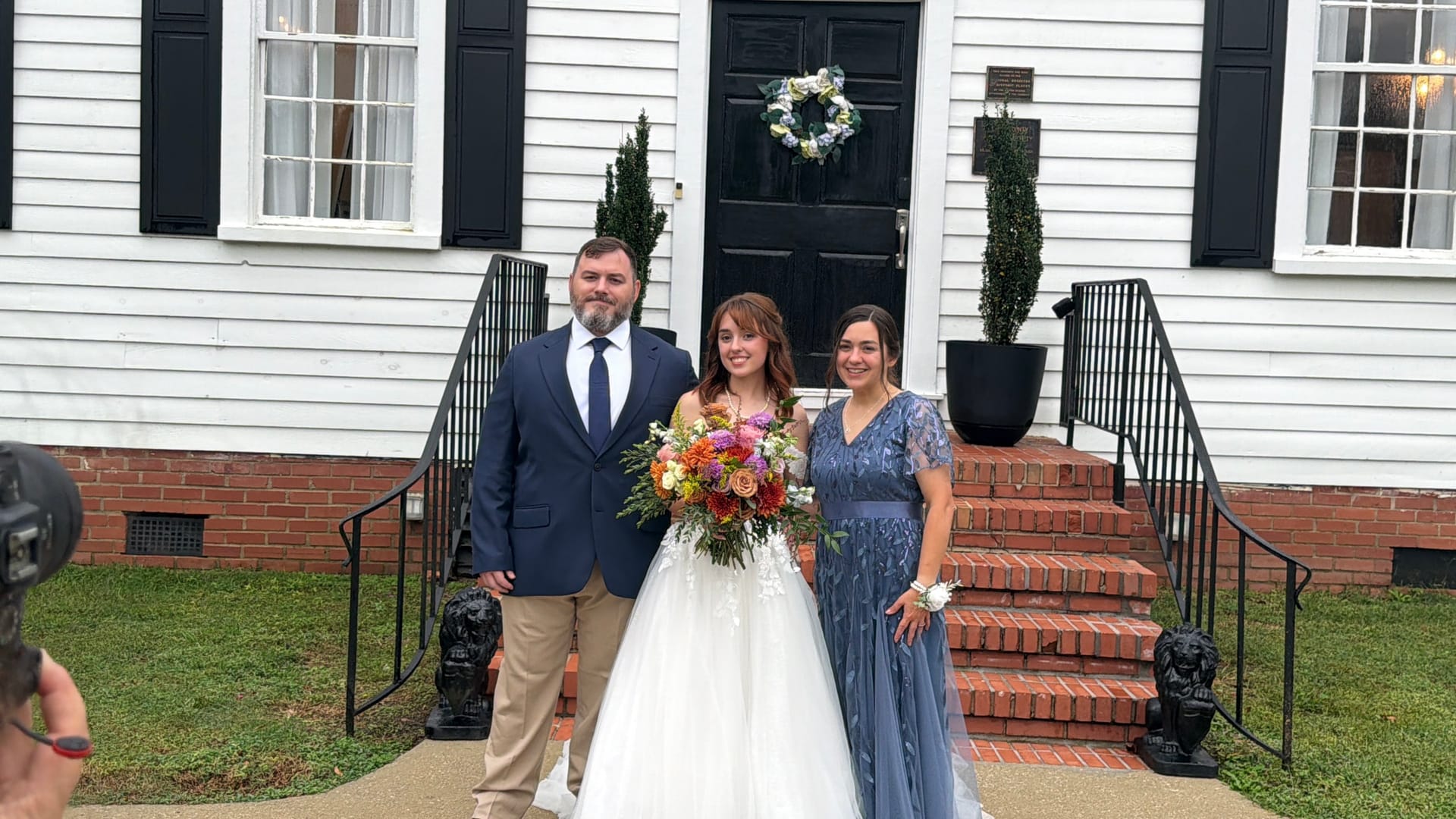 Bride in a white ball gown holding a colorful bridal bouquet stands between two family members on the brick front steps of the 1821 Scarborough House manor.