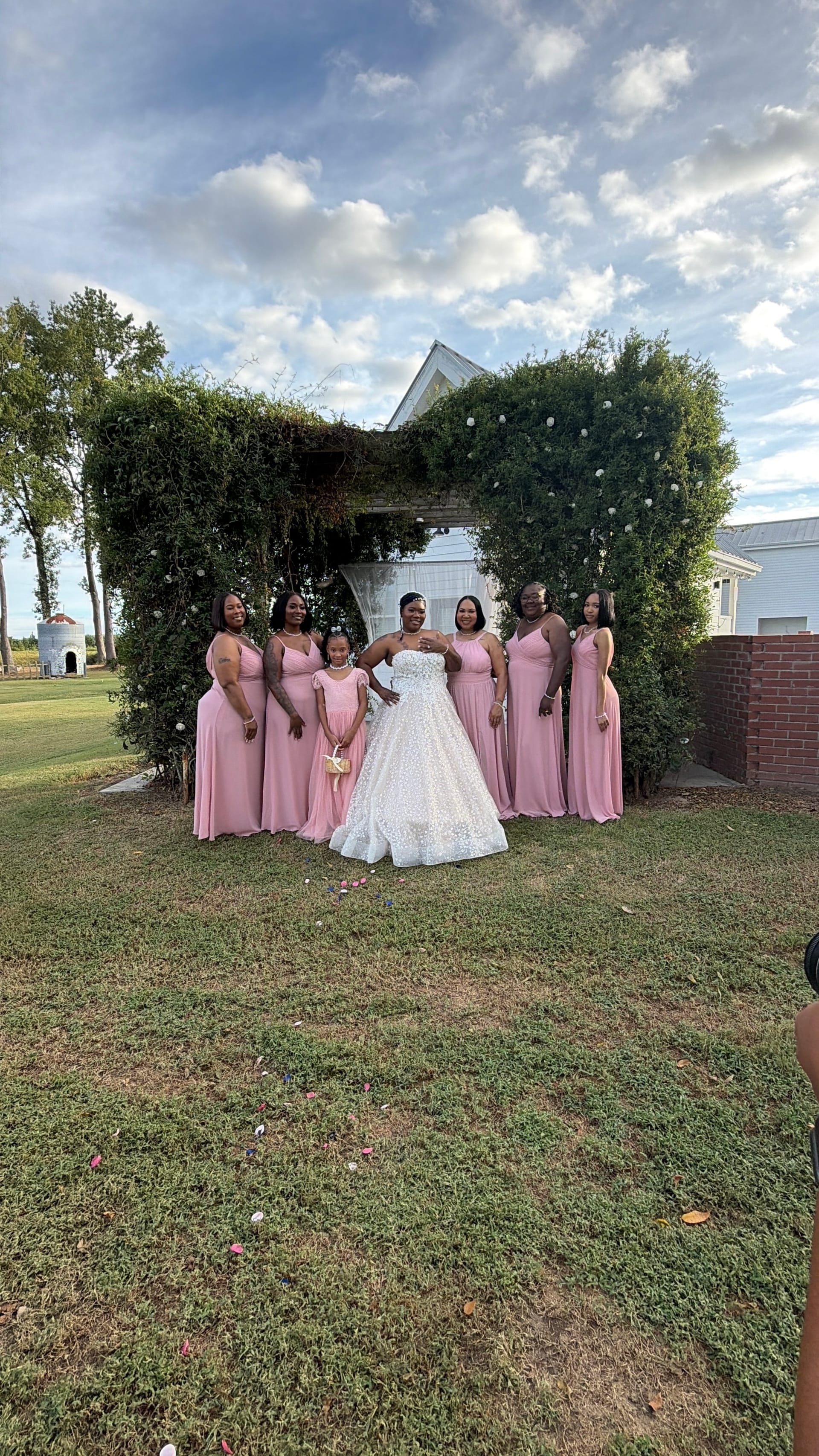 Bride in a sparkling white ball gown poses with her bridesmaids in dusty-rose gowns and a young flower girl in front of a vine-covered archway on the ceremony lawn at Scarborough House.