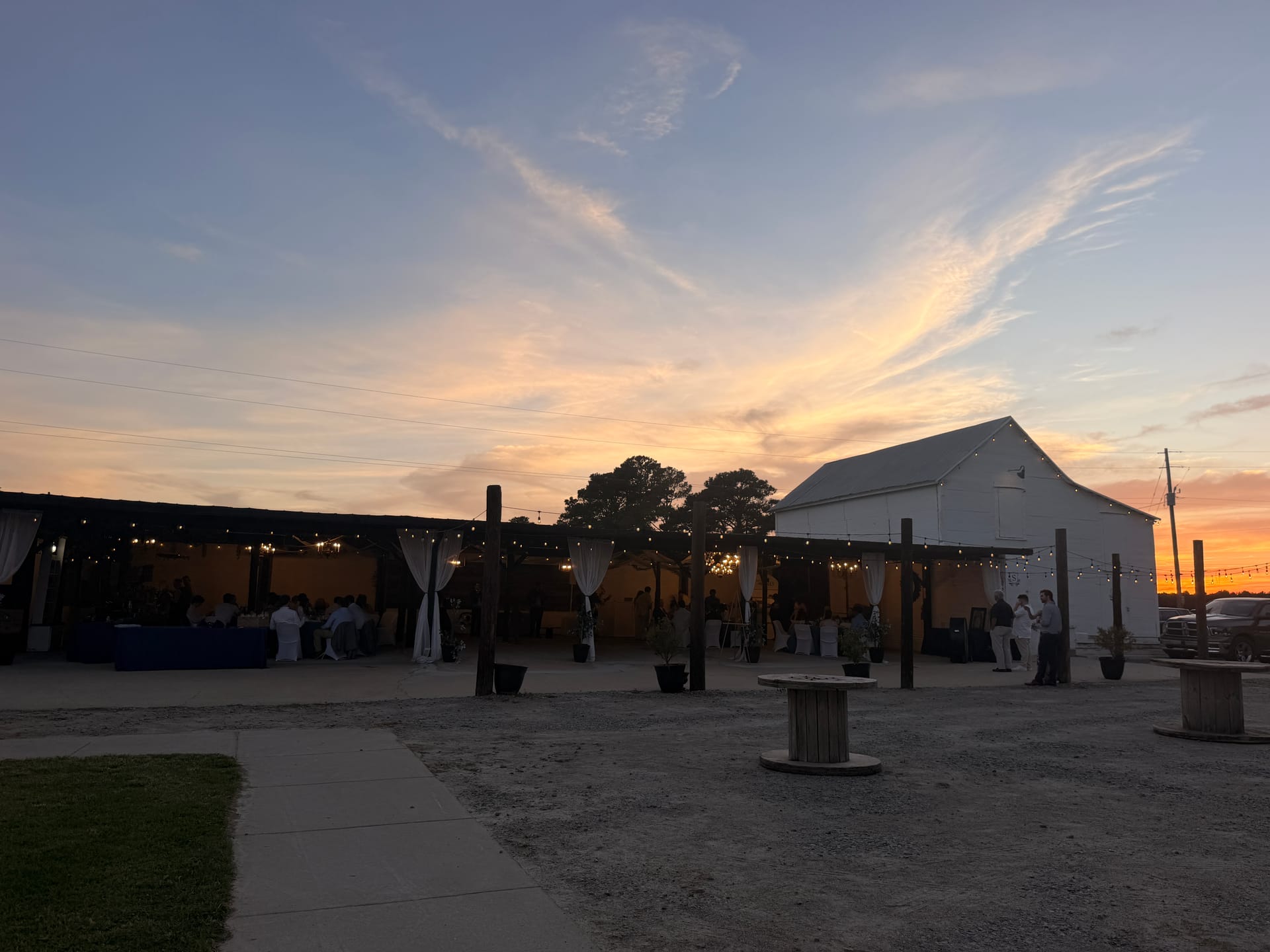 Outdoor wedding reception at dusk with string lights glowing under an open-air pavilion beside a white barn at Scarborough House, set against a vivid orange and blue sunset sky.