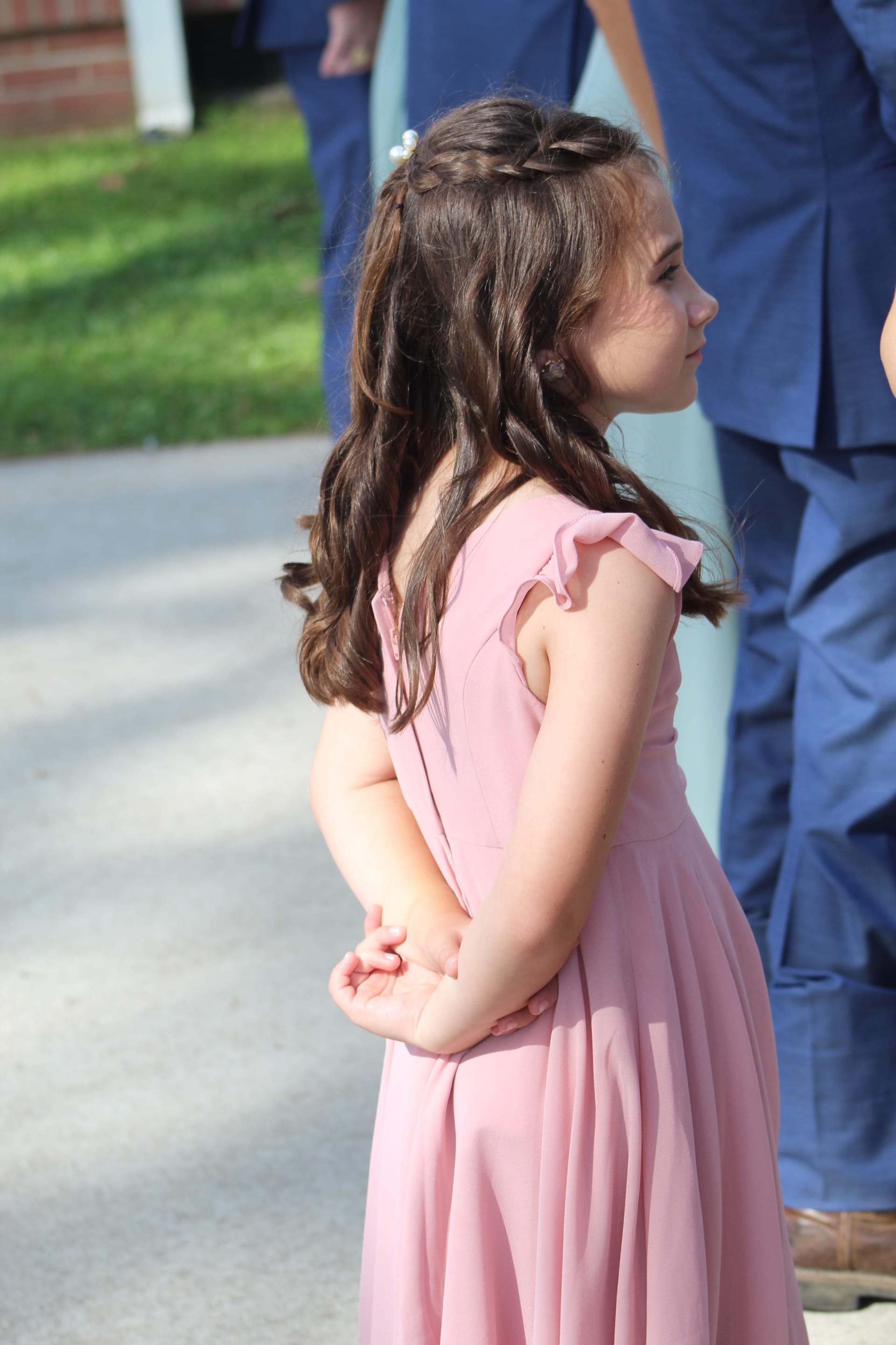 A young flower girl in a blush pink dress with braided hair stands with her hands clasped behind her back during an outdoor wedding ceremony.