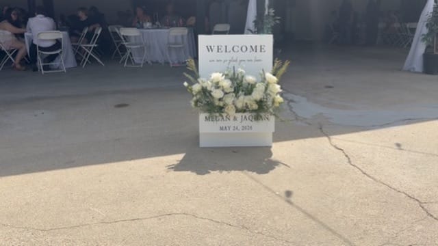 White welcome sign adorned with white roses and greenery announcing the wedding of Megan and Jaquan on May 24 2026 at an outdoor reception venue.