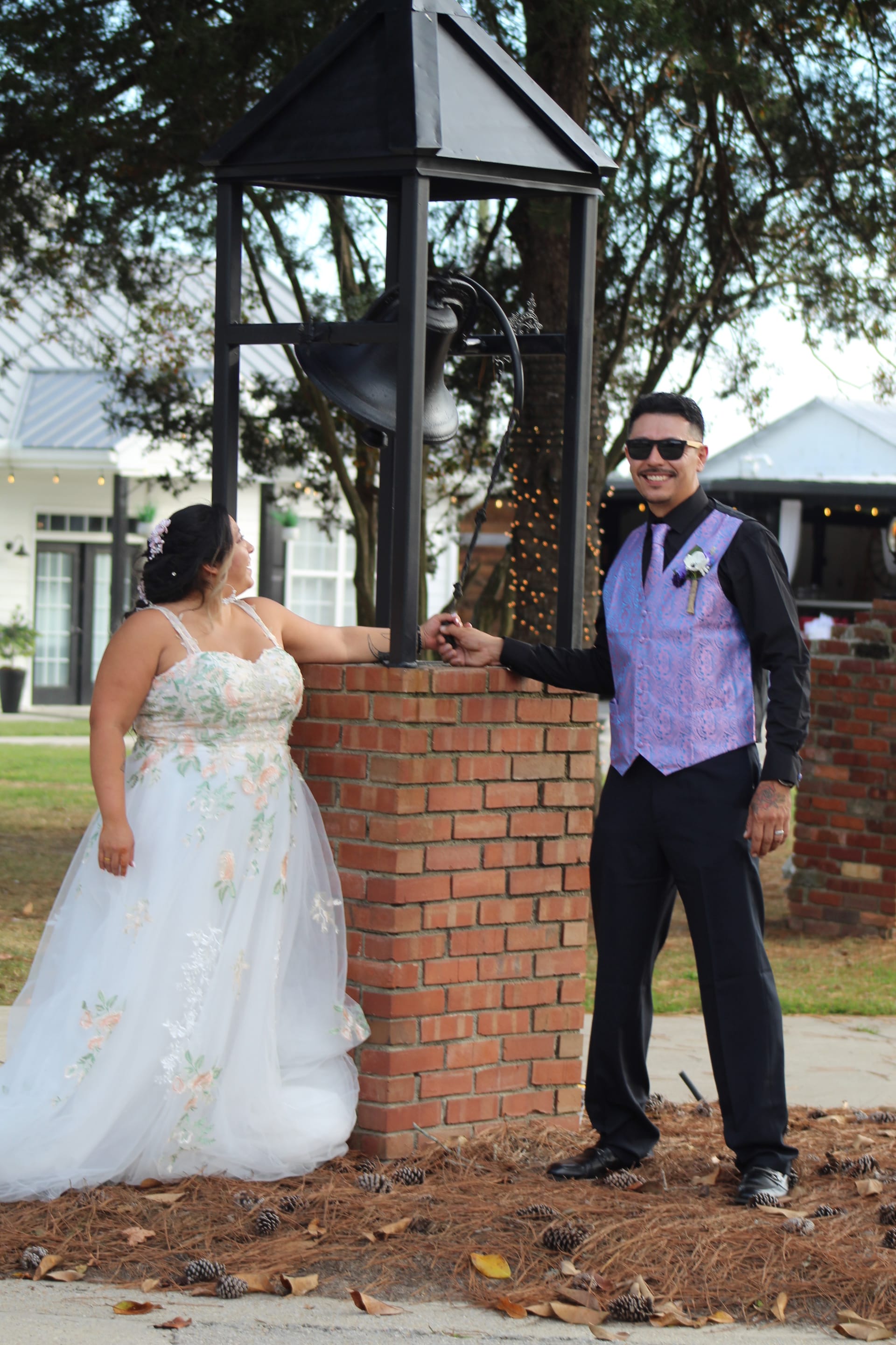 Bride in a floral ballgown and groom in a purple vest and sunglasses hold hands around a brick bell tower at Scarborough House wedding venue.