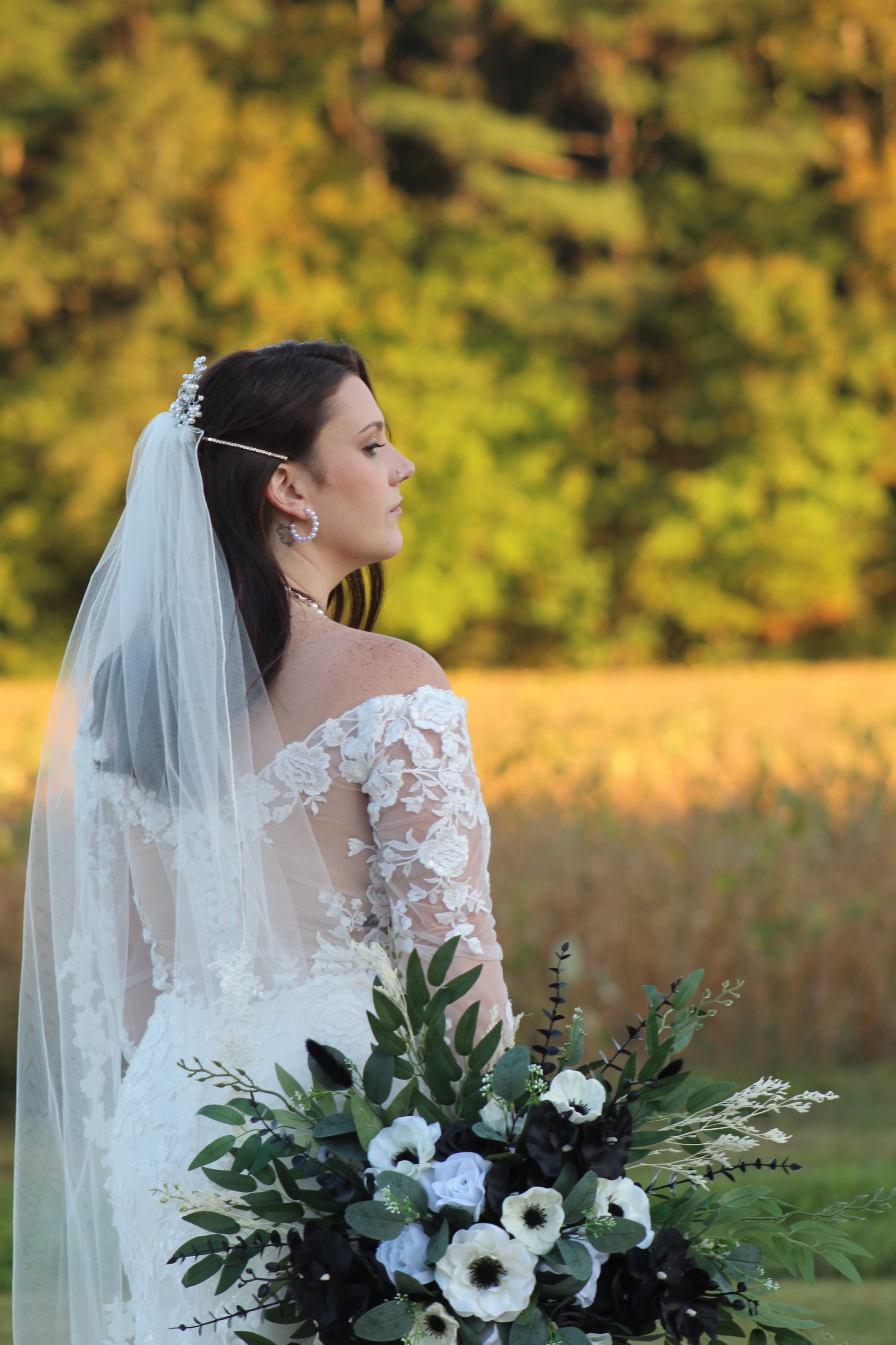 Bride in a lace off-shoulder gown and flowing veil holds a lush black and white floral bouquet while gazing across golden farmland fields at Scarborough House during golden hour.
