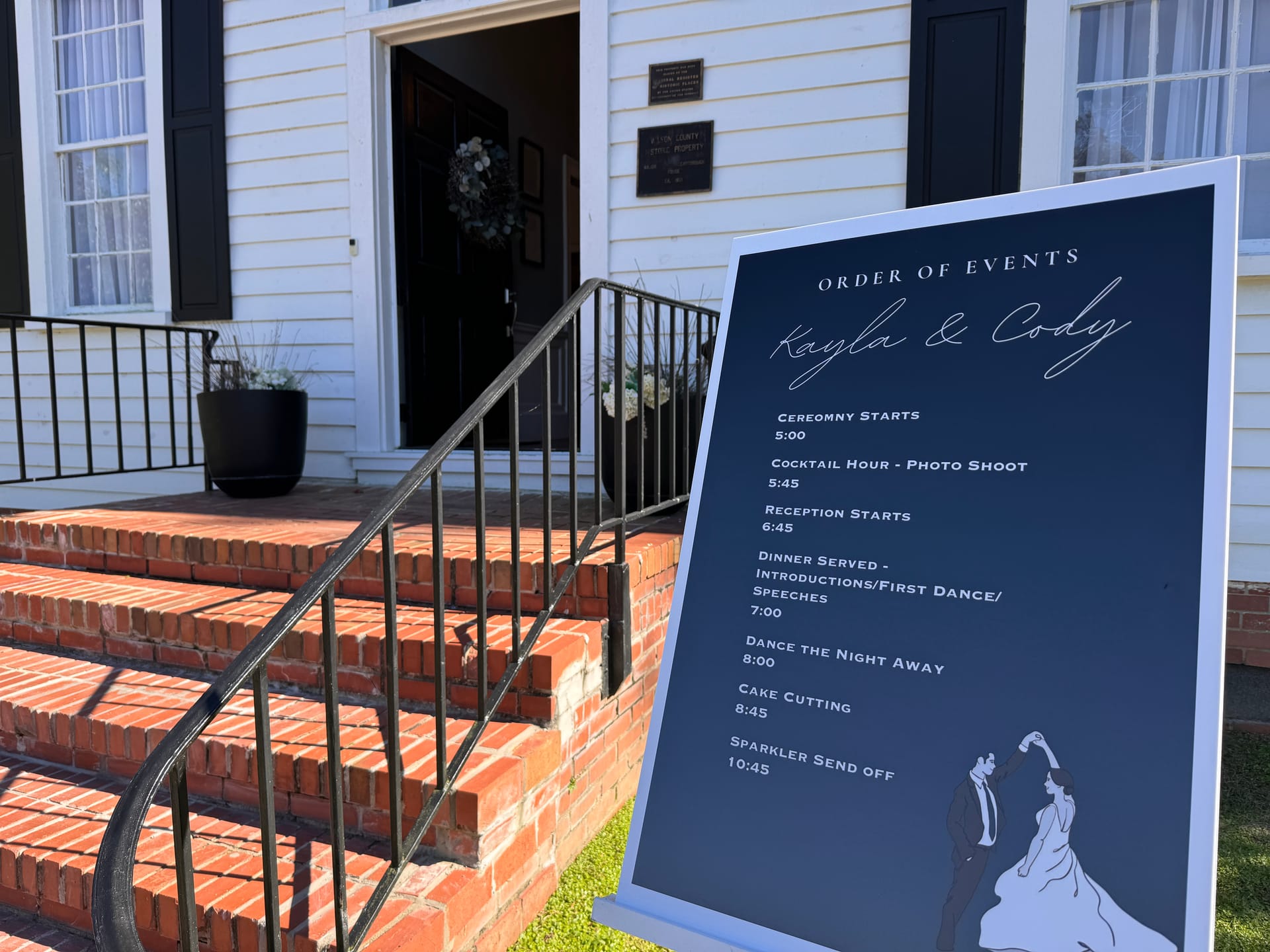 Navy and white Order of Events wedding sign for Kayla and Cody displayed on the brick front steps of the 1821 Scarborough House historic manor entrance.