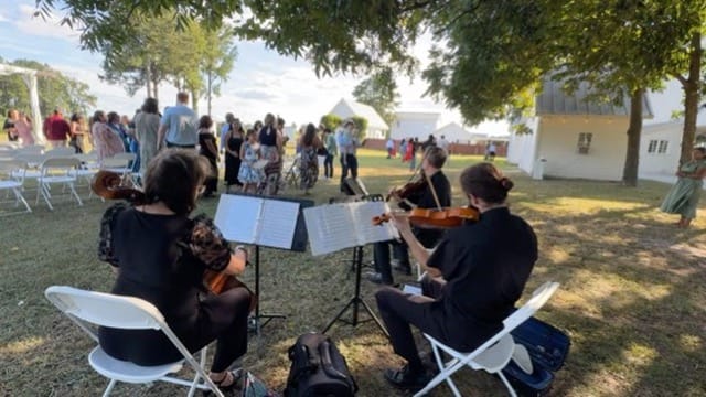 A string quartet performs outdoors on the ceremony lawn at Scarborough House as wedding guests mingle in the background beneath oak trees near the historic white manor.