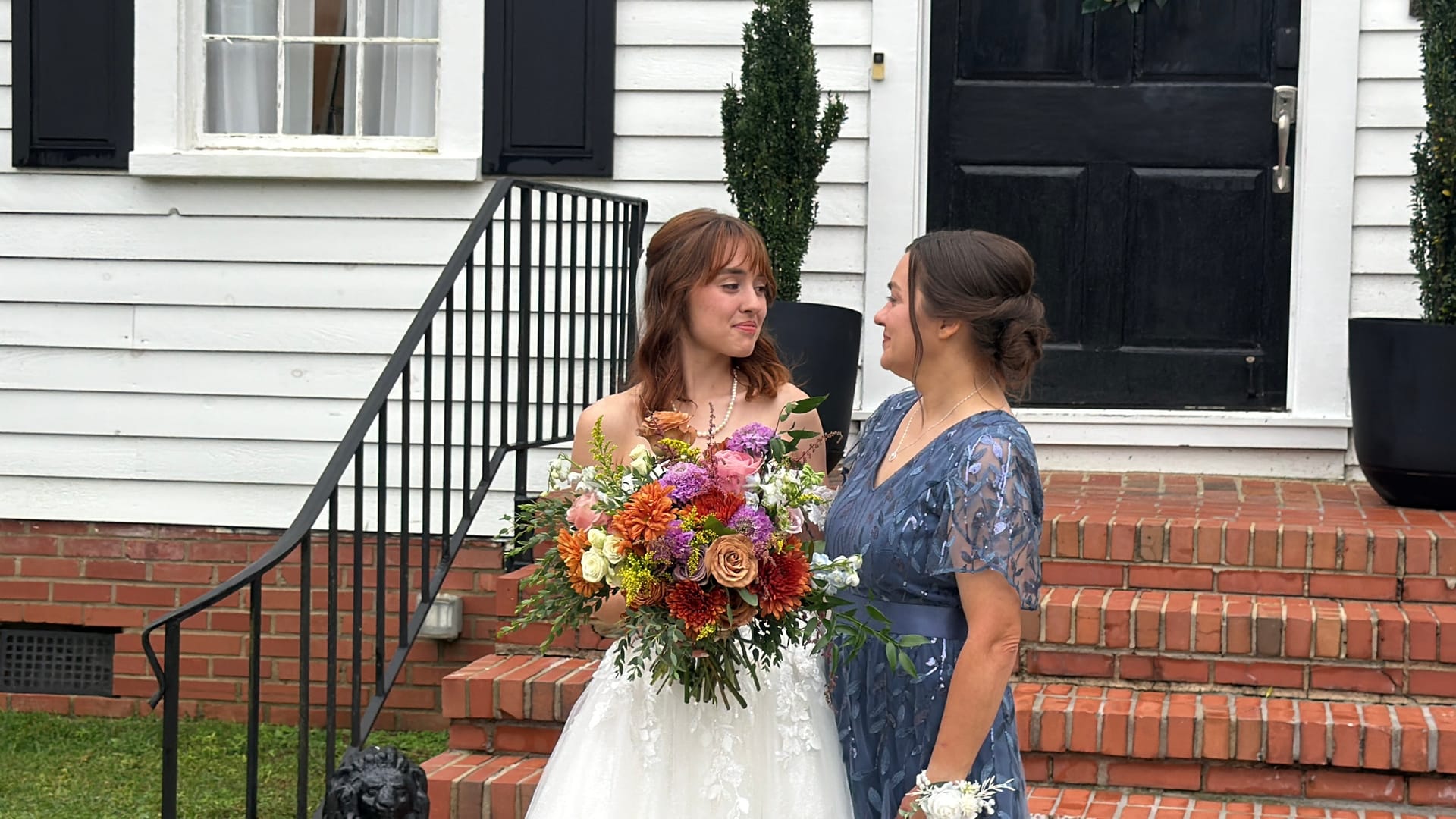 Bride in a white lace gown holding a lush colorful bouquet shares a warm glance with a woman in a blue dress on the brick front steps of the historic 1821 Scarborough House manor.
