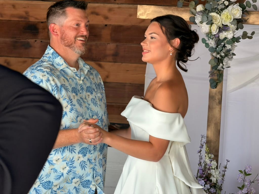 Bride in an off-shoulder white gown and groom in a floral shirt share a joyful first dance beneath a rustic wood-paneled backdrop adorned with white and lavender floral arrangements at Scarborough House.