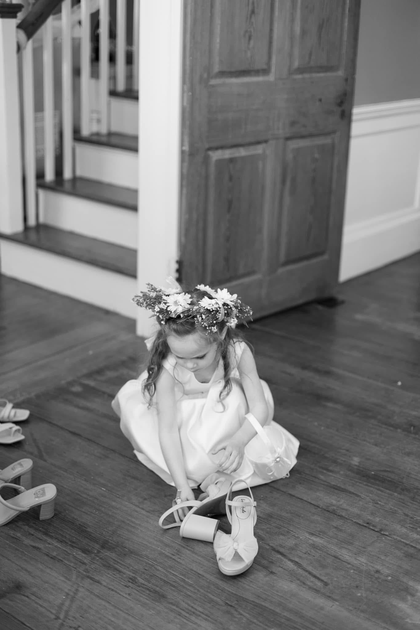 Flower girl in a white dress and floral crown sits on a hardwood floor putting on her heeled sandals inside the historic Scarborough House manor.