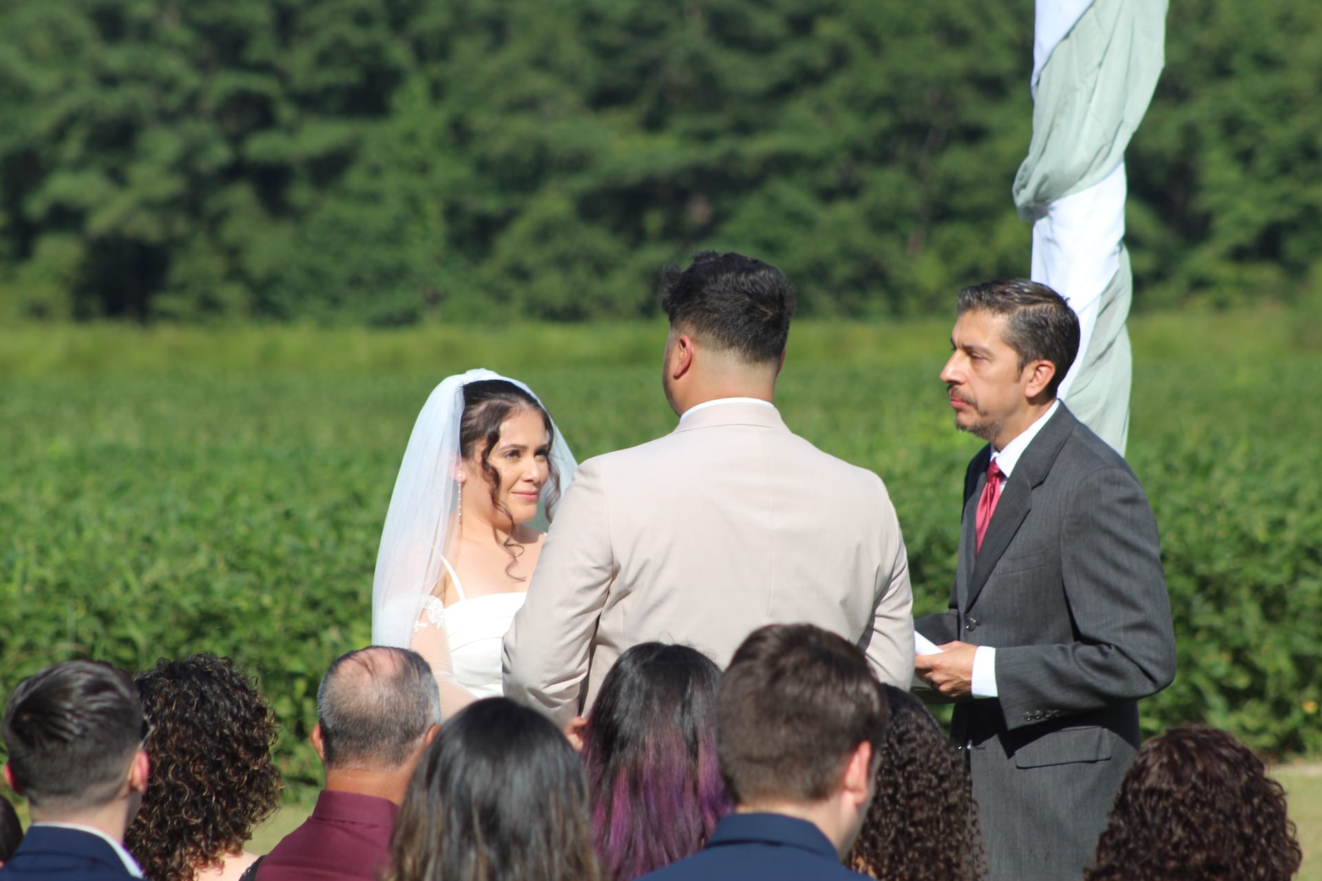 Bride in white gown and veil exchanges vows with groom as officiant looks on during an outdoor ceremony at Scarborough House with green farmland fields and trees in the background.