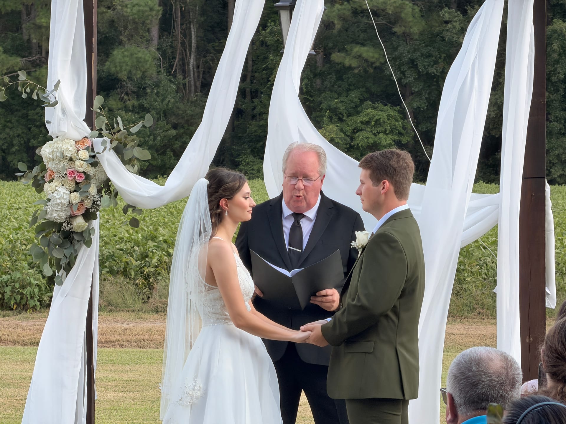 Bride and groom hold hands at an elegant outdoor altar adorned with white draping and a lush floral arrangement while an officiant reads from a book at Scarborough House wedding venue.