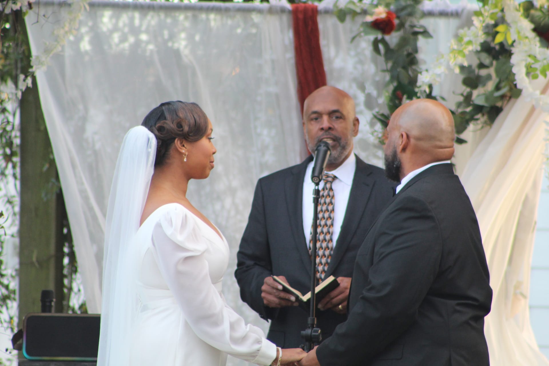 A bride in a white gown and veil faces her groom as an officiant reads from a book at a microphone beneath a decorated floral arch during an outdoor wedding ceremony.