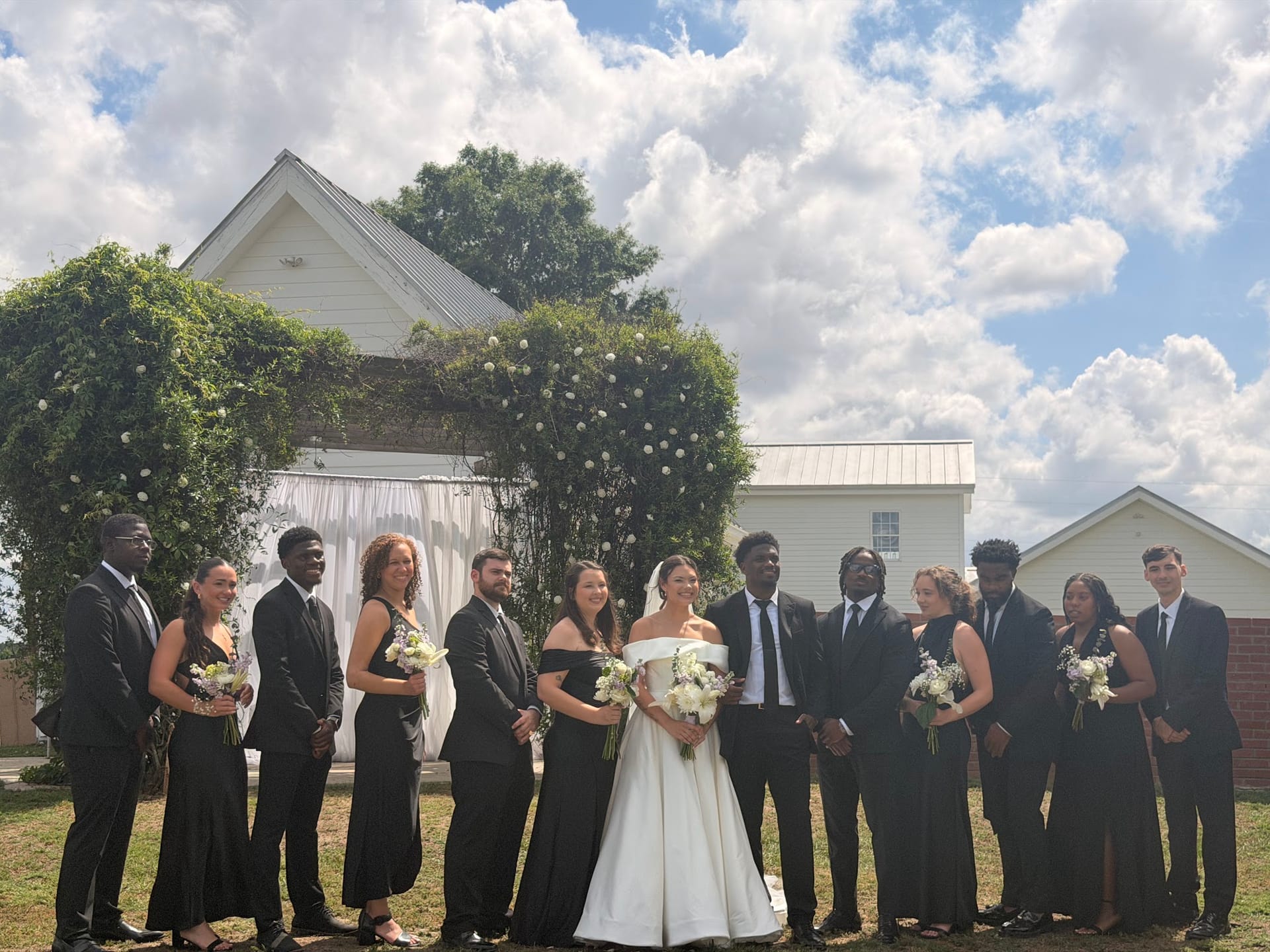 Bride and groom surrounded by their wedding party in black formal attire posing in front of a white ivy-draped chapel arch at Scarborough House under a dramatic cloudy sky.