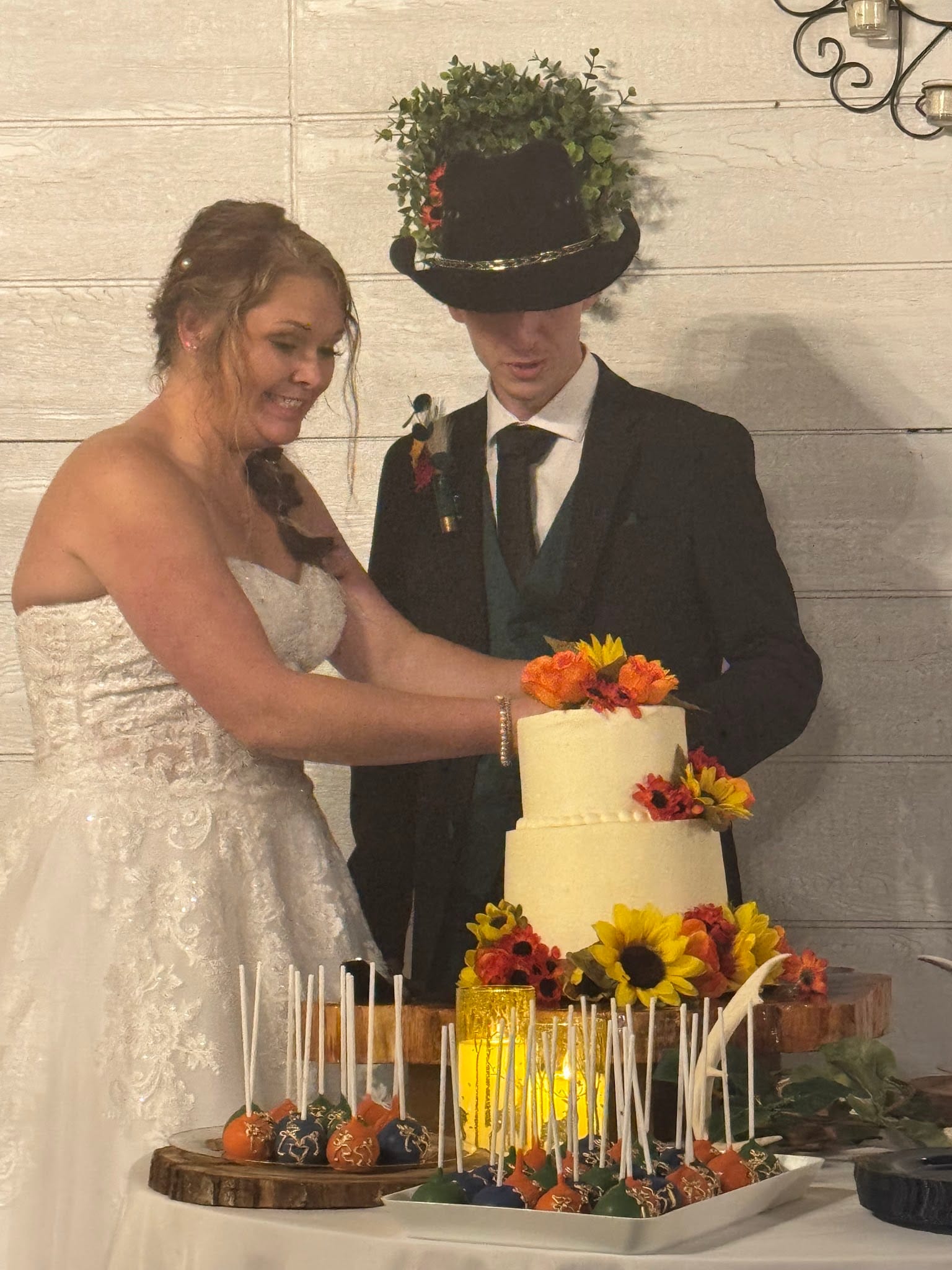 Bride and groom cutting a two-tier white wedding cake adorned with sunflowers and autumn flowers at Scarborough House, surrounded by colorful cake pops on a rustic wood display.