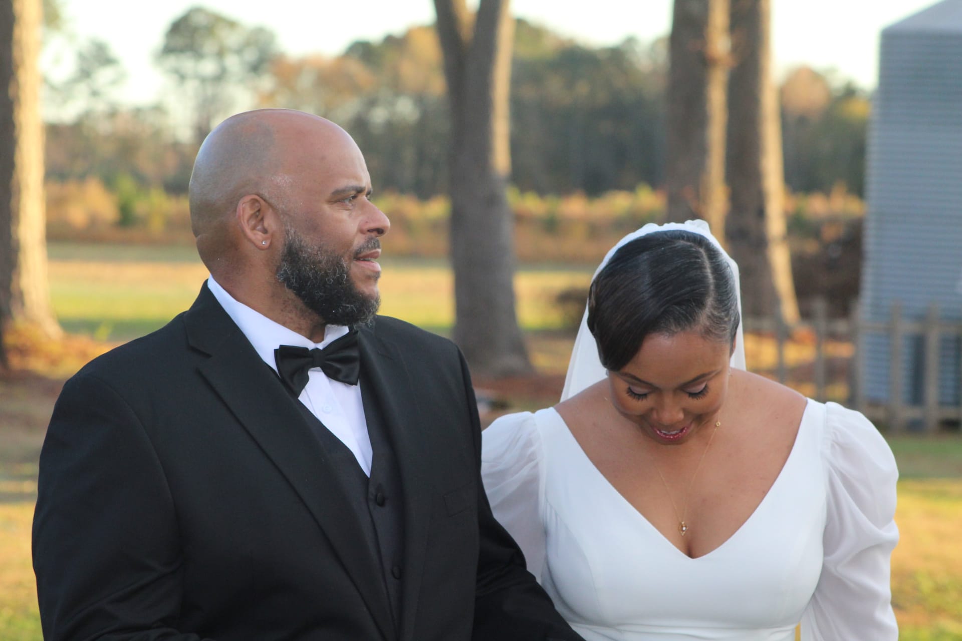 Groom in black tuxedo and bow tie gazes tenderly at his bride in a white gown and veil during golden-hour portraits on the farmland grounds of Scarborough House estate.