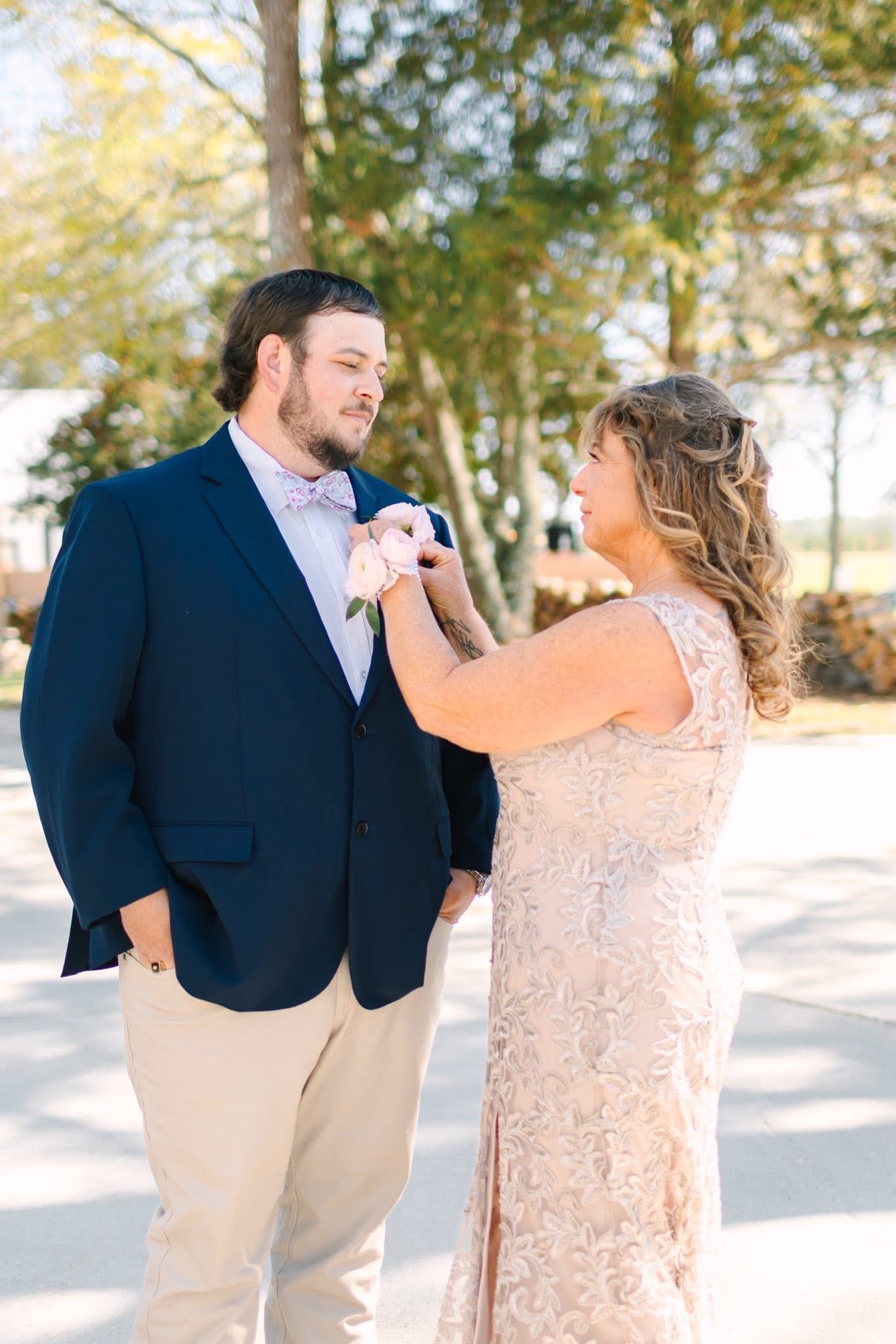 A woman in a blush lace gown pins a pink boutonniere onto a groom in a navy suit and floral bow tie outdoors at a sunlit wedding venue.