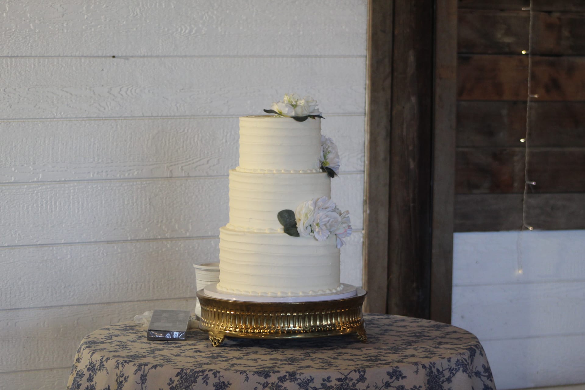 Elegant three-tier white buttercream wedding cake adorned with white florals and greenery displayed on a gold cake stand at Scarborough House historic estate venue.