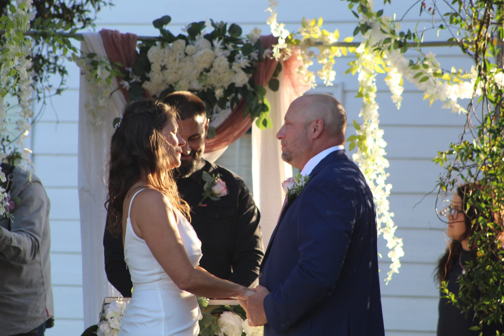 Bride and groom hold hands and exchange vows beneath a white floral and greenery wedding arch at Scarborough House outdoor ceremony lawn.