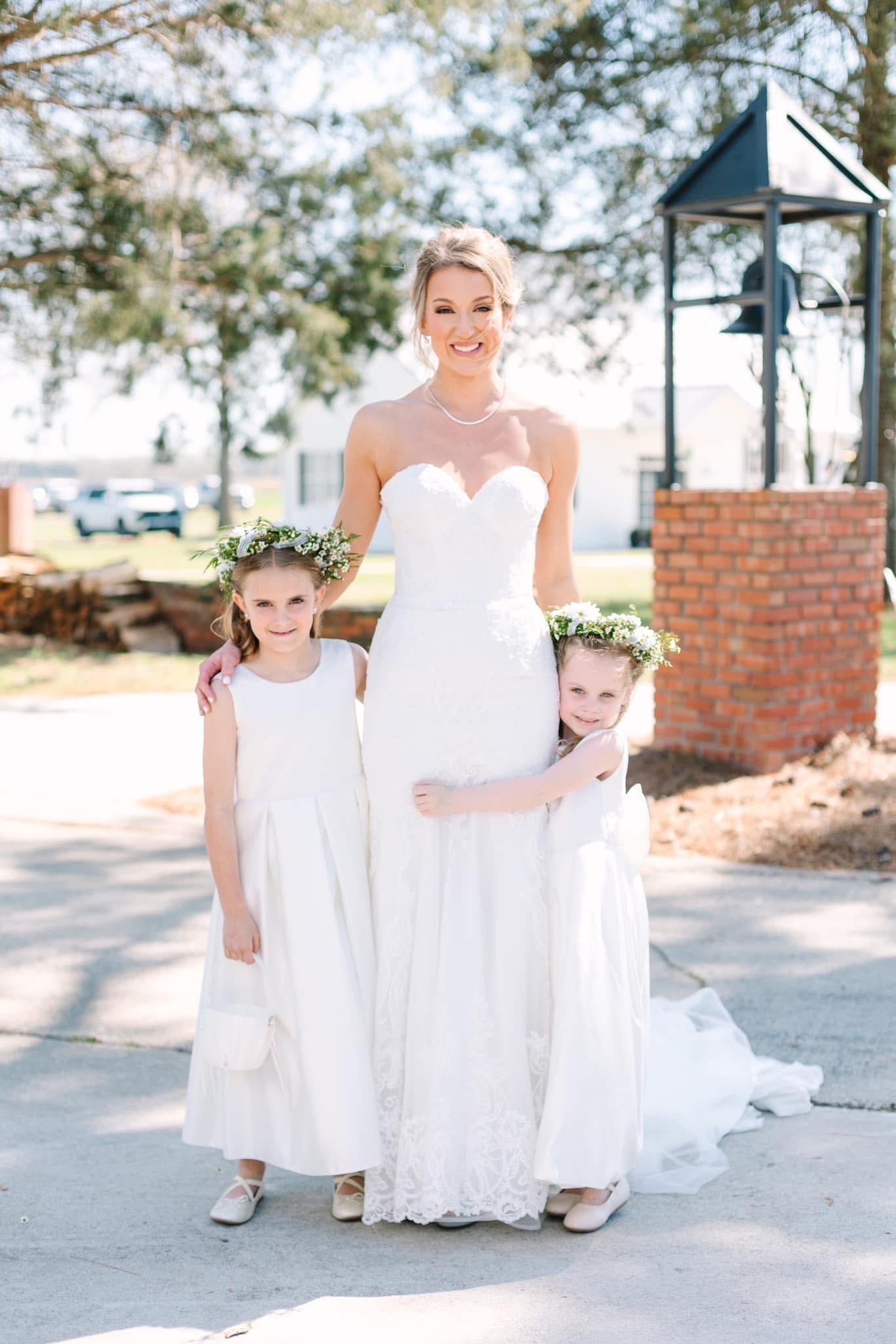 Smiling bride in a strapless lace gown poses with two flower girls wearing floral crowns and white dresses near the historic bell and brick pillar at Scarborough House.