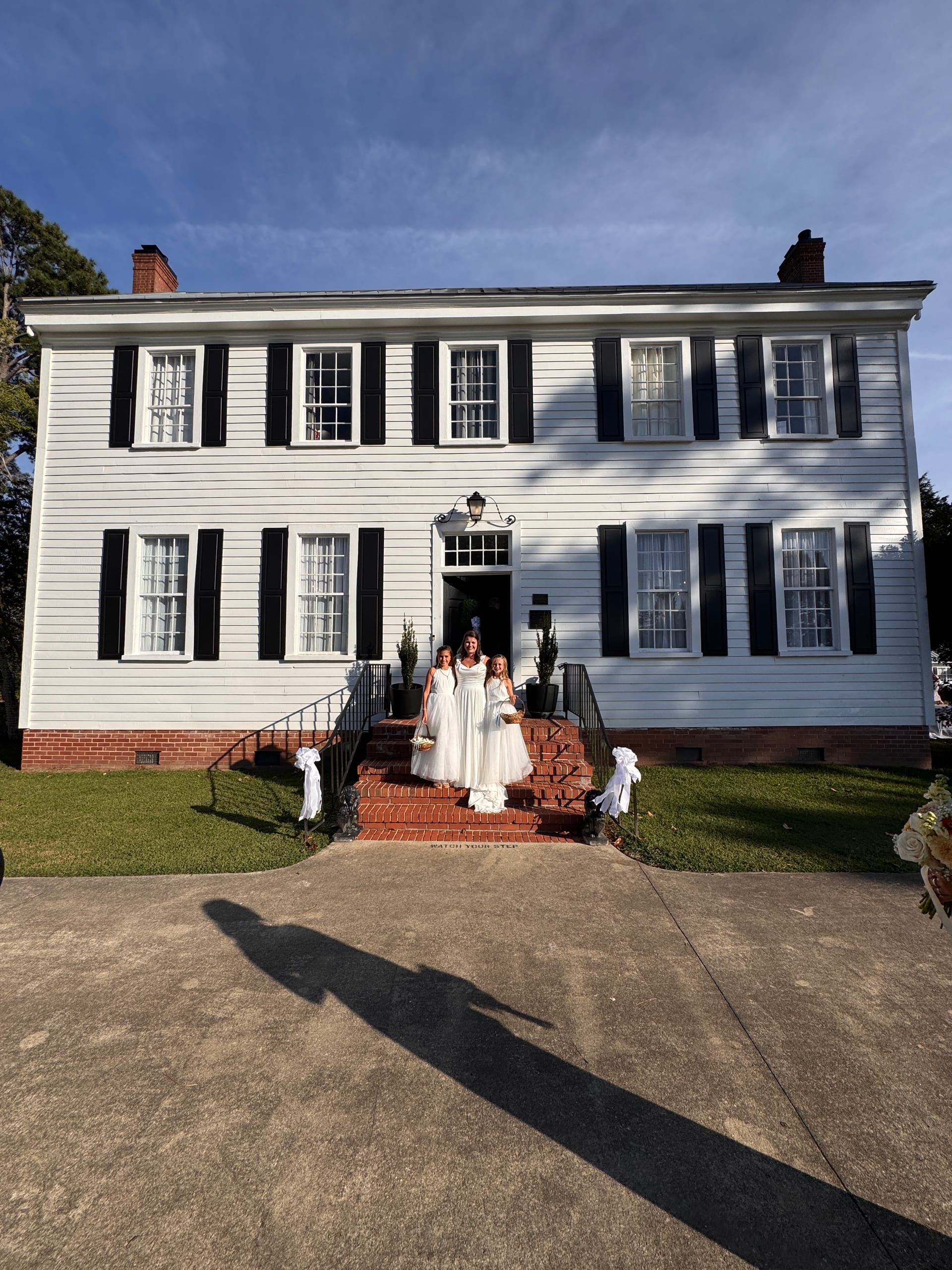 Bride in a flowing white gown stands on the brick front steps of the 1821 Scarborough House manor flanked by two young flower girls in white dresses, with the classic white colonial facade and black shutters framing the scene.