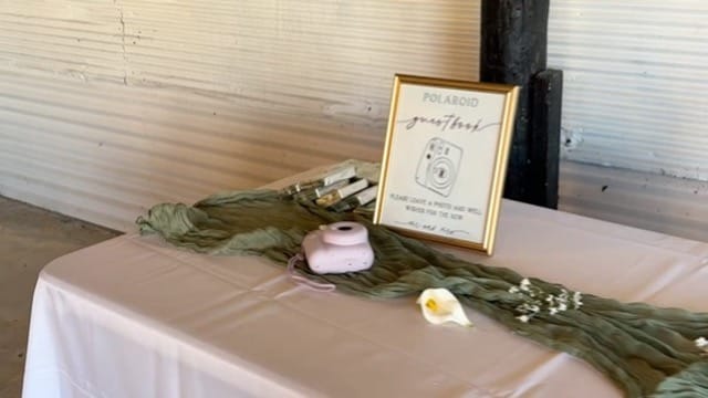 A styled wedding guestbook table draped with a sage green cheesecloth runner featuring a pink Instax camera, white calla lily, and gold-framed Polaroid guestbook sign.