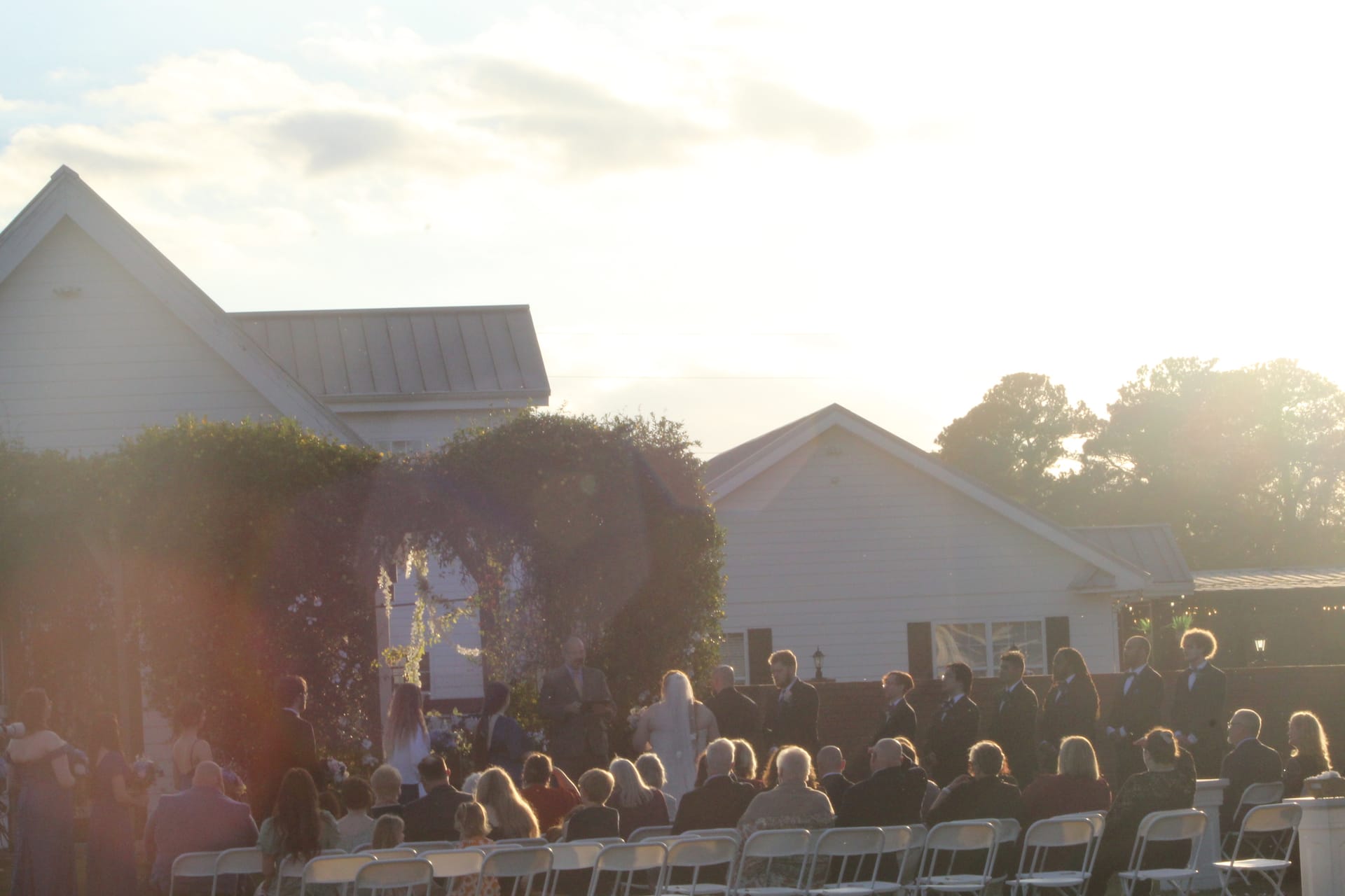 Guests seated in white chairs watch a wedding ceremony at golden hour beneath a lush floral arch on the ceremony lawn at Scarborough House historic estate.