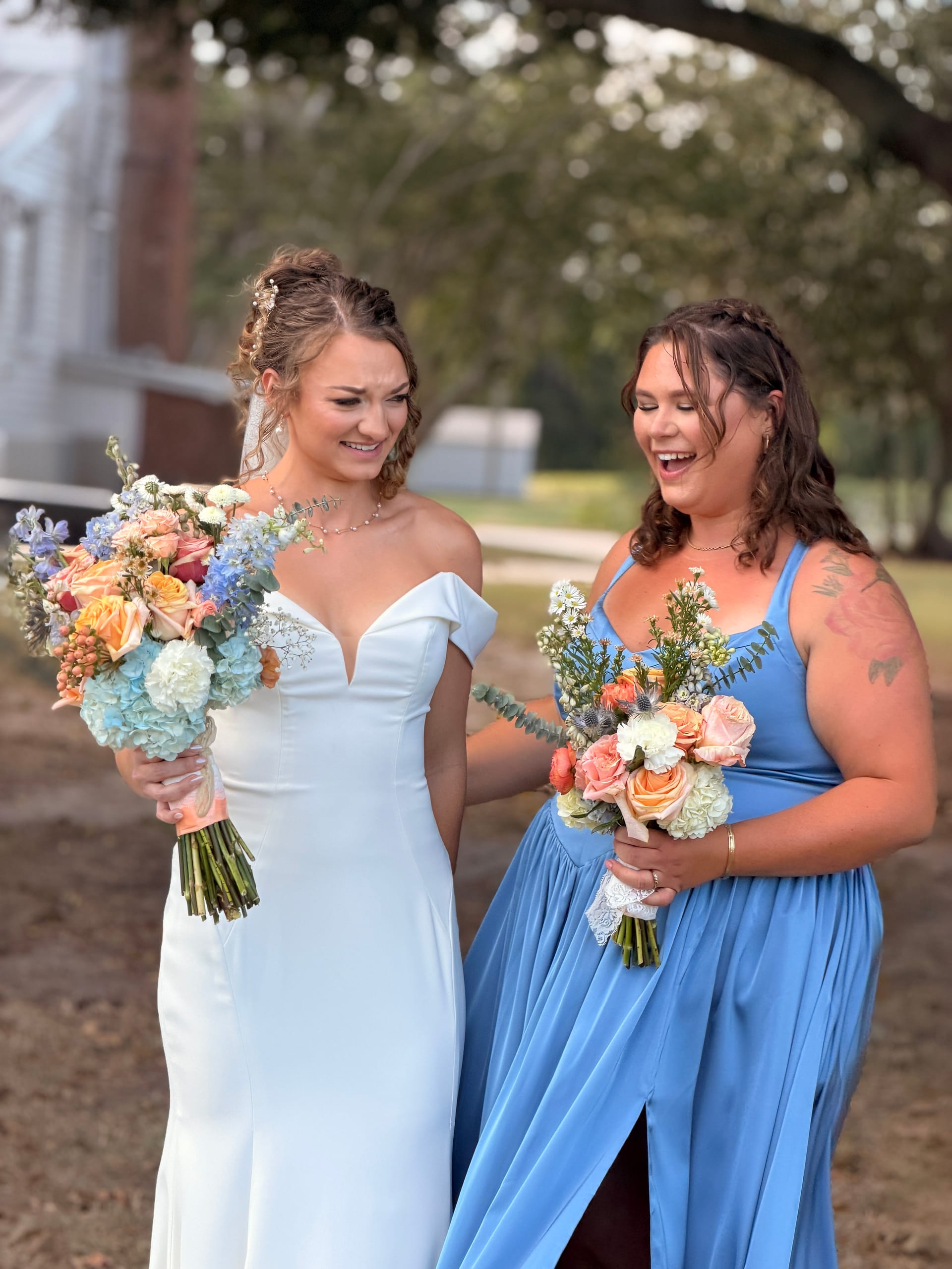Bride in white off-shoulder gown and bridesmaid in cornflower blue dress share a joyful laugh while holding colorful peach and blue floral bouquets beneath sweeping oak trees at Scarborough House.