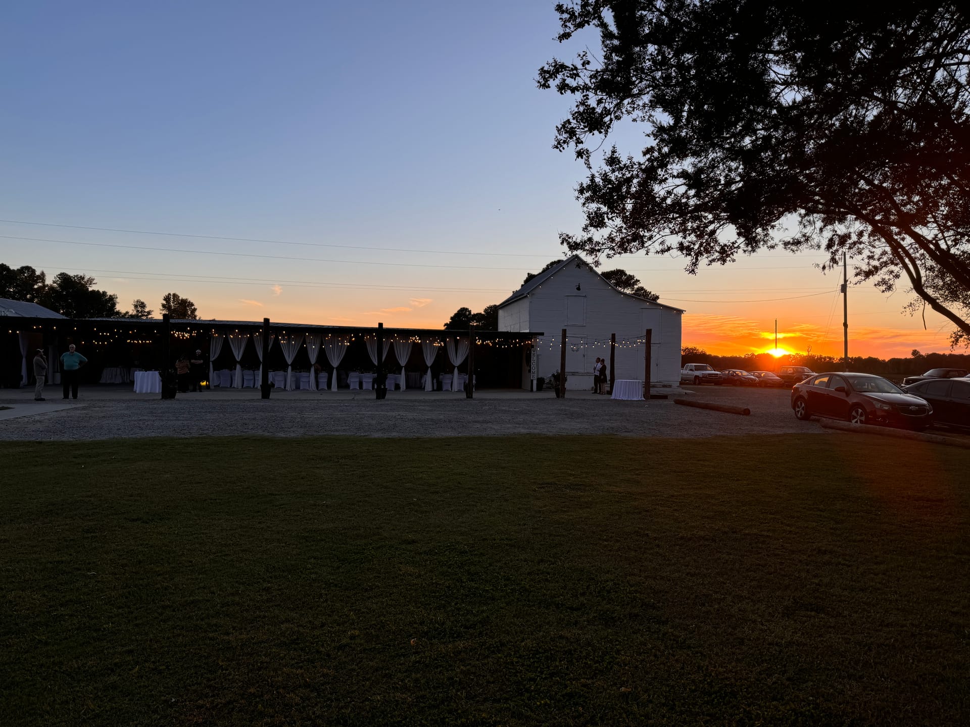 Silhouette of a white barn wedding venue with string lights and draped curtains glowing against a vibrant orange and blue sunset sky at Scarborough House.