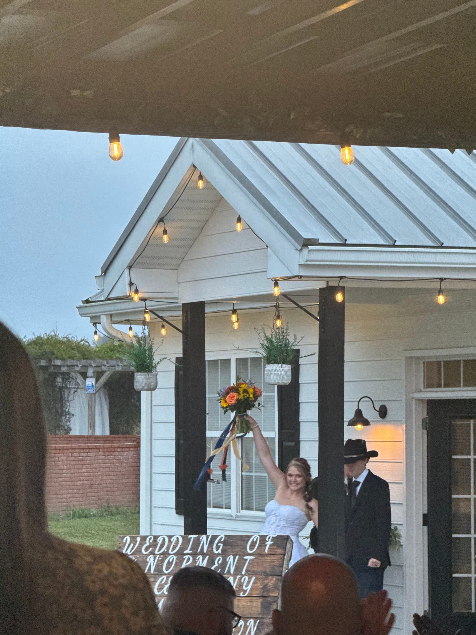 Bride in white strapless gown triumphantly raises her colorful bouquet beside her groom in a black cowboy hat outside the illuminated white farmhouse at Scarborough House, framed by warm string lights at dusk.