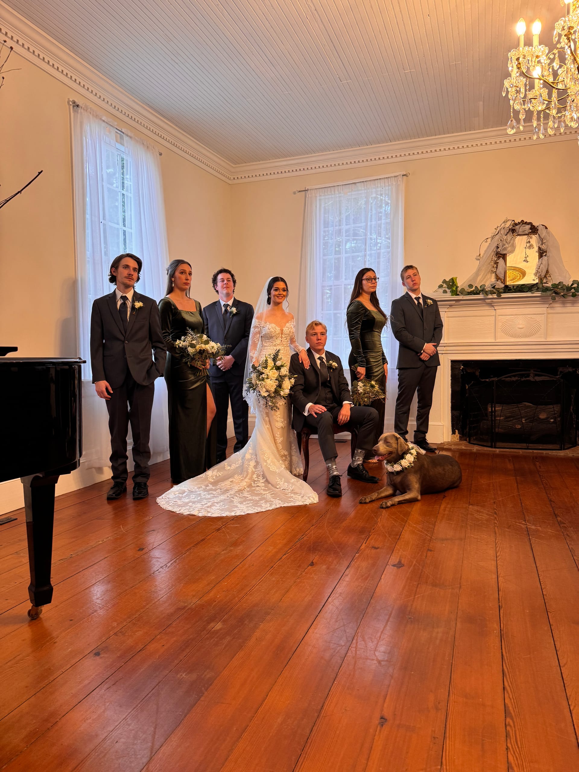 Bride in lace gown and groom seated with their wedding party in dark formal attire posing beside a fireplace in the grand hall of the 1821 Scarborough House estate, accompanied by a flower-crowned dog.