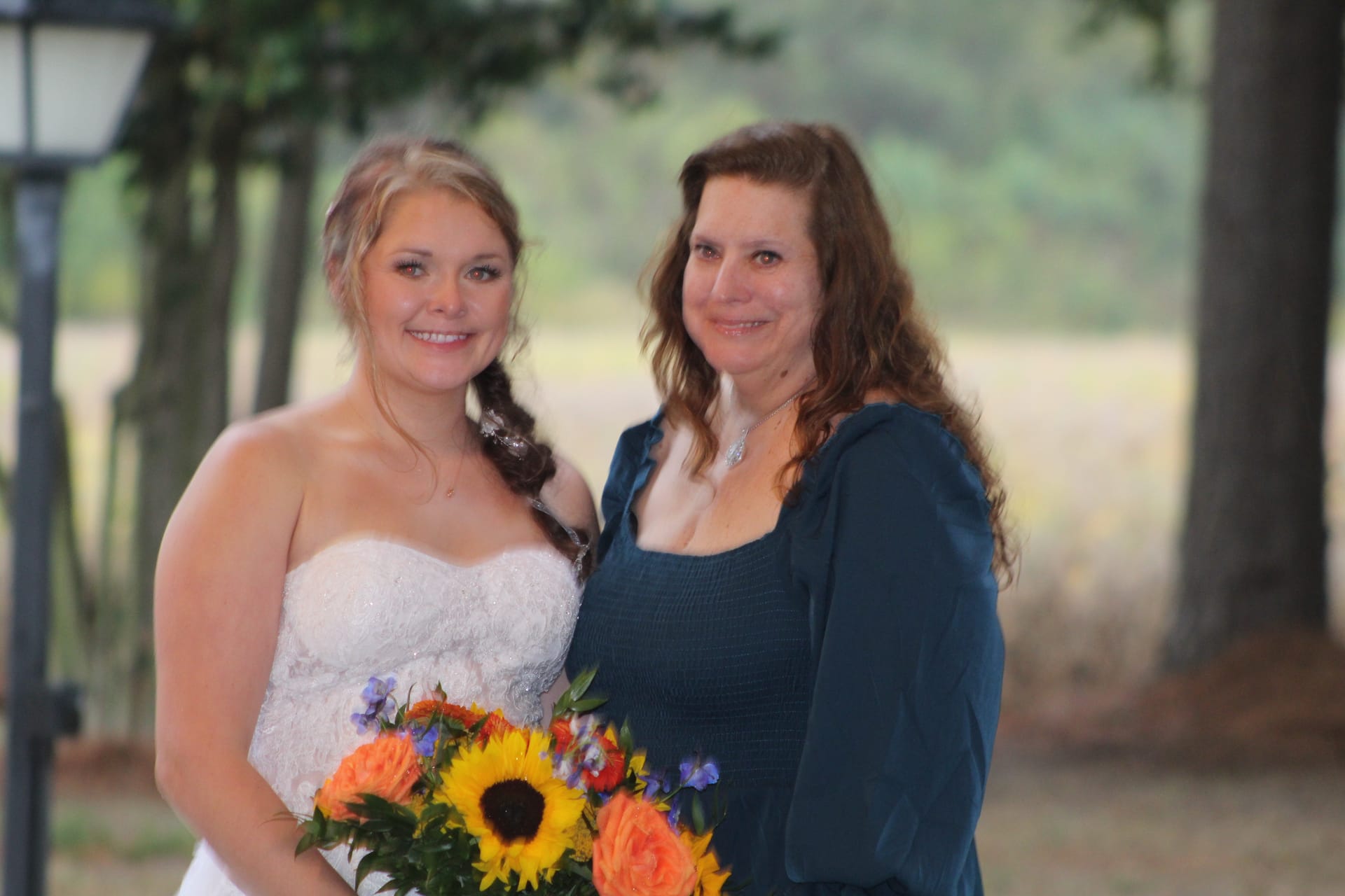 Bride in a strapless lace gown holding a sunflower and orange rose bouquet poses outdoors with a woman in teal against a softly blurred oak tree and field backdrop at Scarborough House.