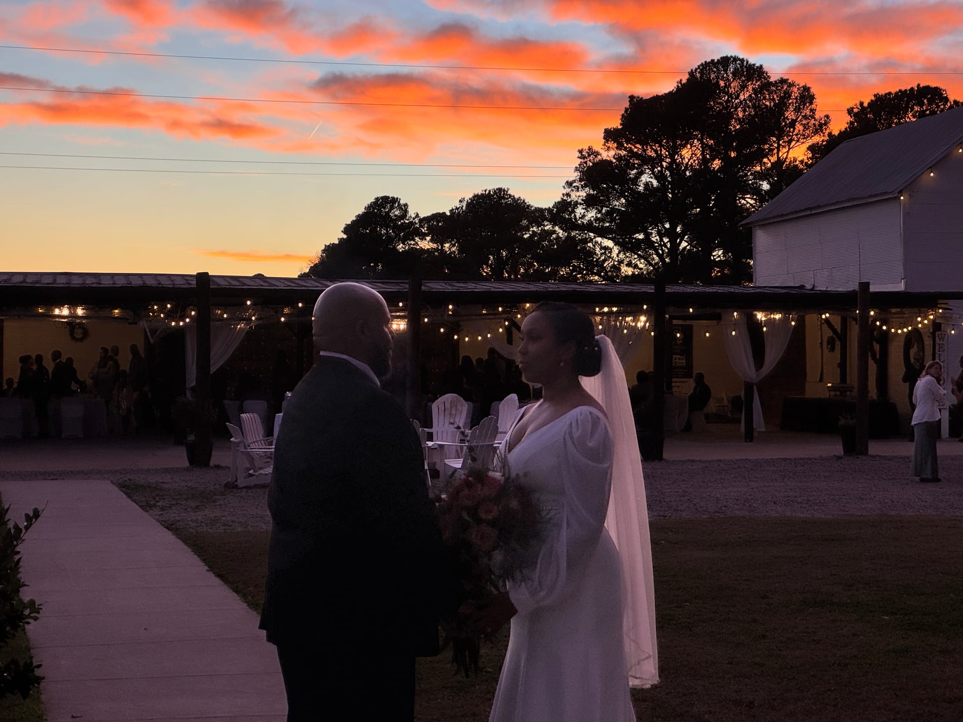 Bride in a white gown holding a bouquet and groom in a dark suit face each other on the ceremony lawn at Scarborough House beneath a breathtaking orange and pink sunset sky with string lights glowing behind them.