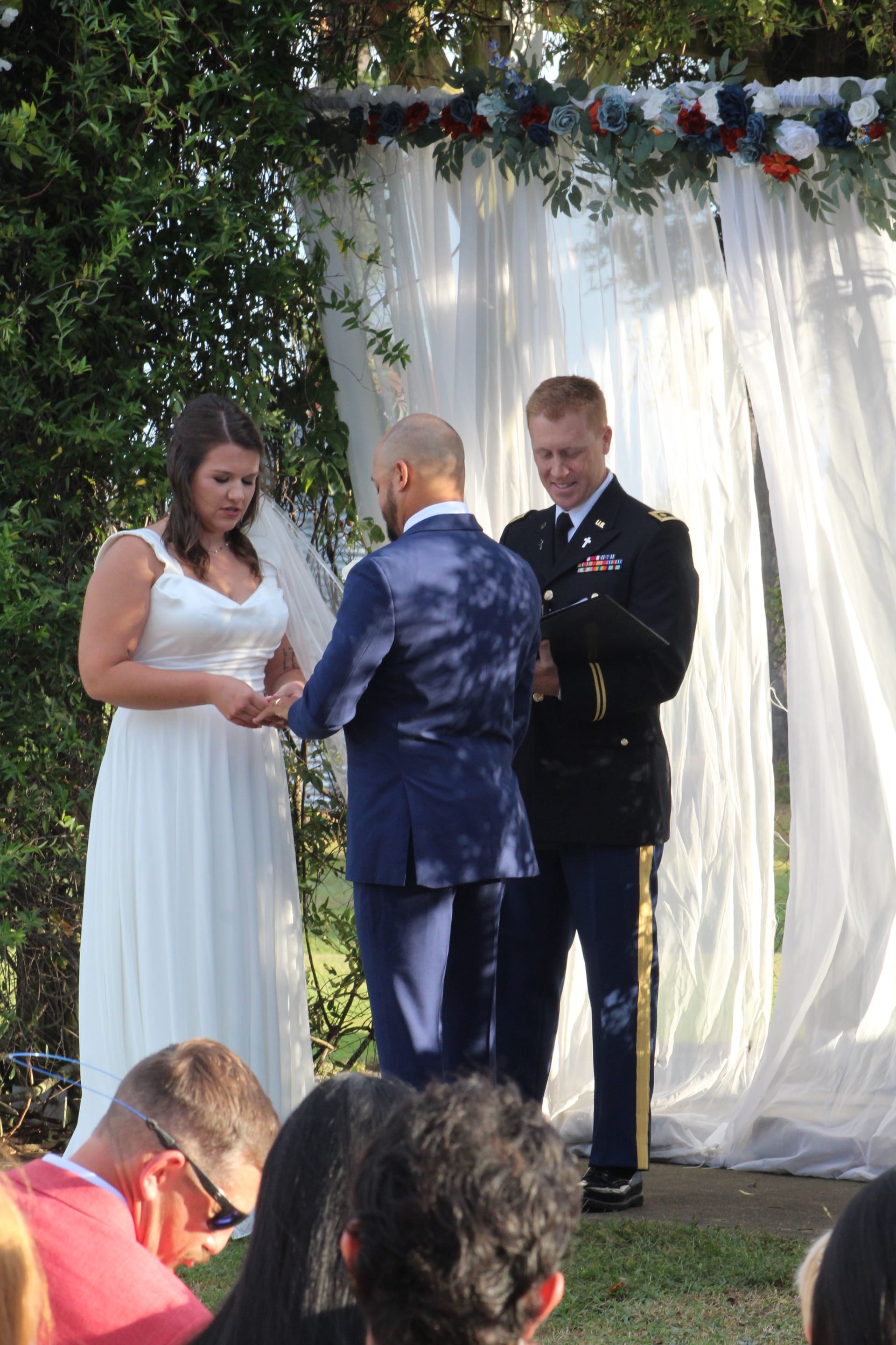 Bride and groom exchange rings during an outdoor wedding ceremony beneath a draped arch decorated with red white and blue flowers officiated by a military officer.