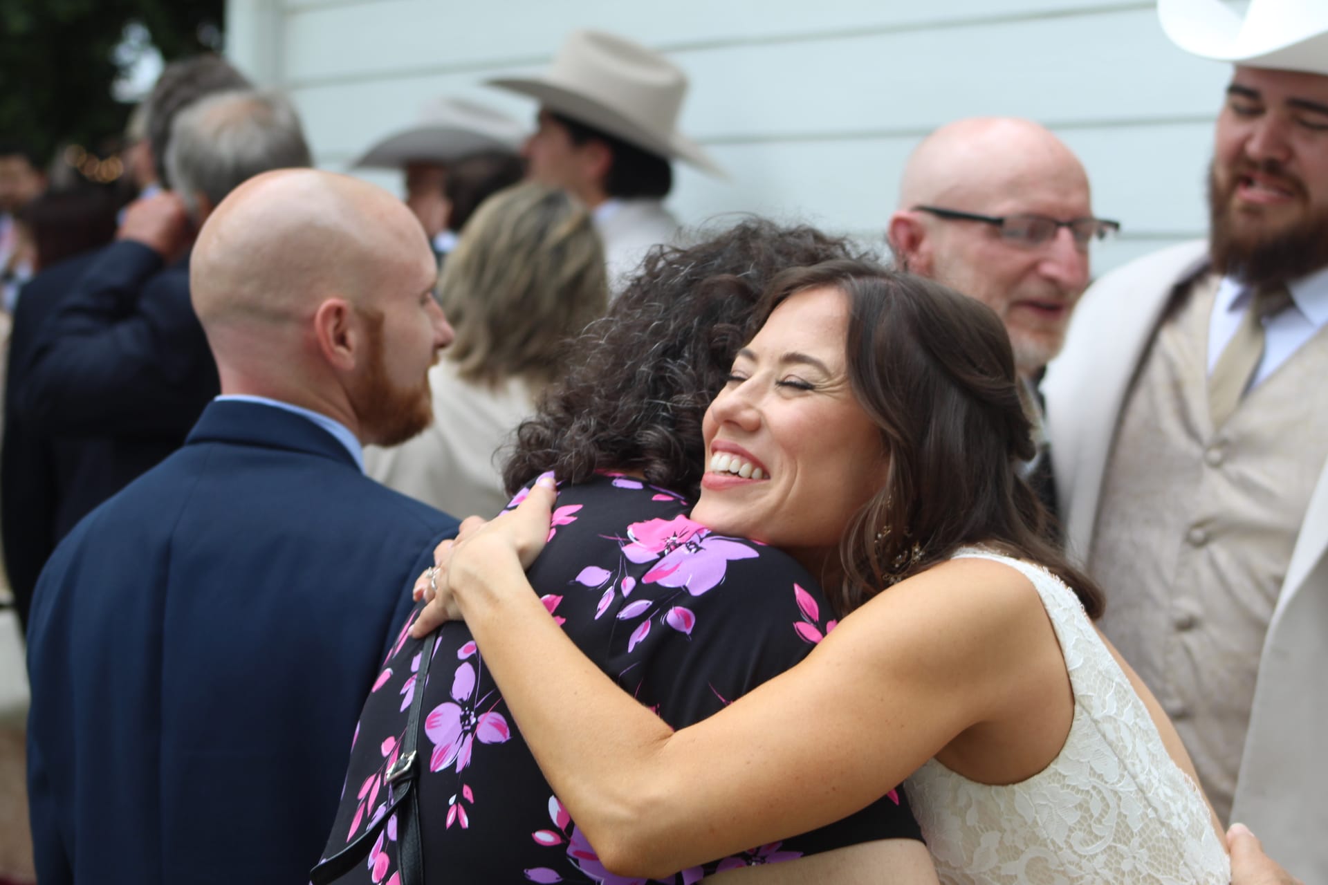 A smiling bride in a white lace dress shares a warm embrace with a guest in a floral dress during an outdoor wedding celebration at Scarborough House.