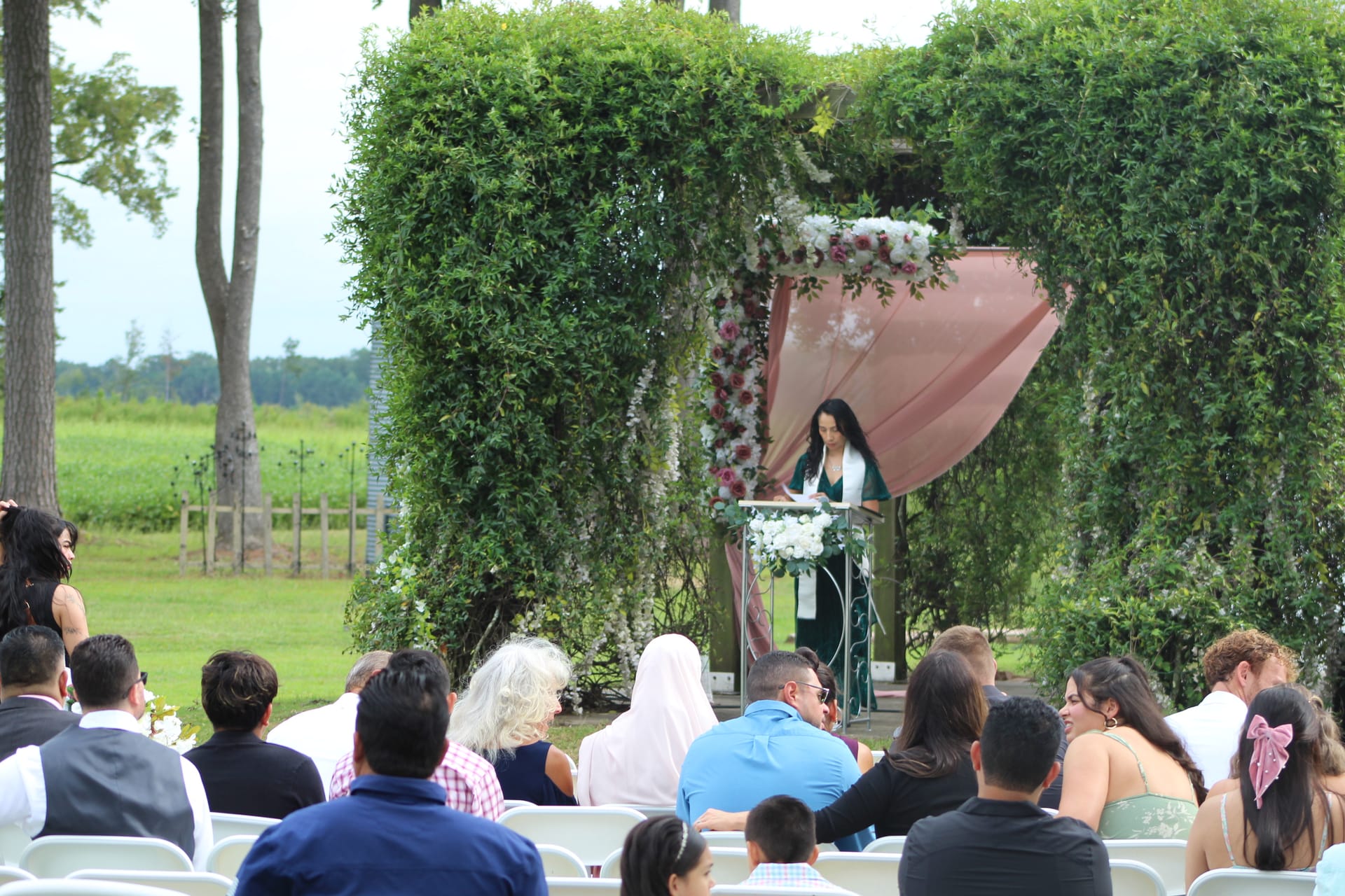 Wedding guests seated on white chairs watching an officiant speak beneath a lush greenery arch adorned with pink draping and floral arrangements on the ceremony lawn at Scarborough House.