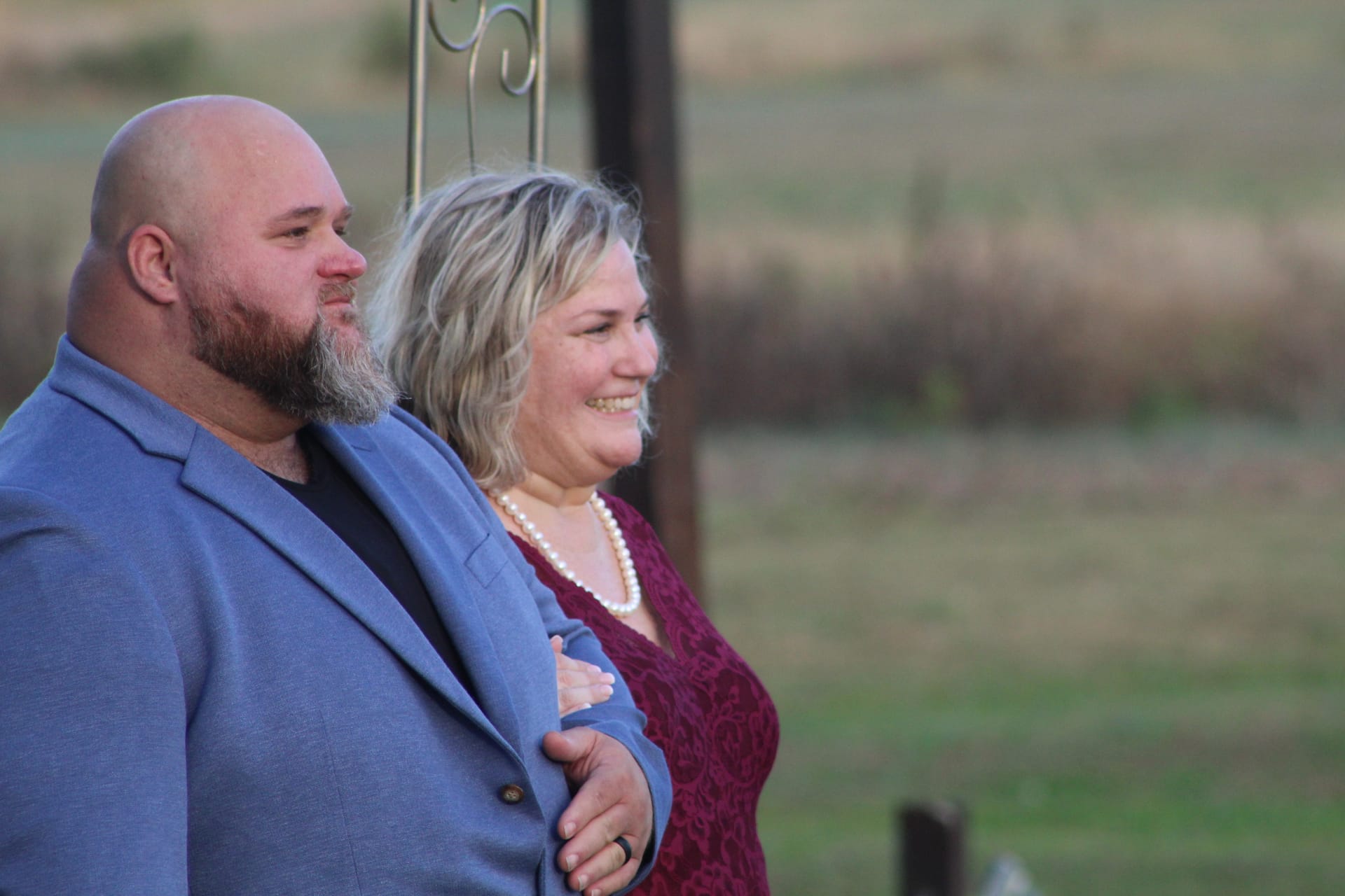 A smiling couple dressed in a blue blazer and burgundy lace dress stand arm-in-arm at golden hour with soft farmland fields in the background at Scarborough House.